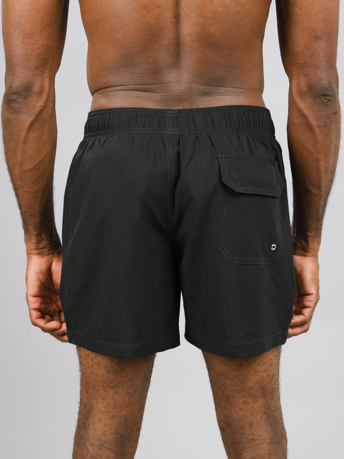 An individual faces away from the camera, donning ID Menswears Oyogu Quick Dry 4 Way Stretch Swim Trunk. These black shorts, crafted from quick-dry recycled plastic, include an elastic waistband and a right-side back pocket. The backdrop is a plain light gray.