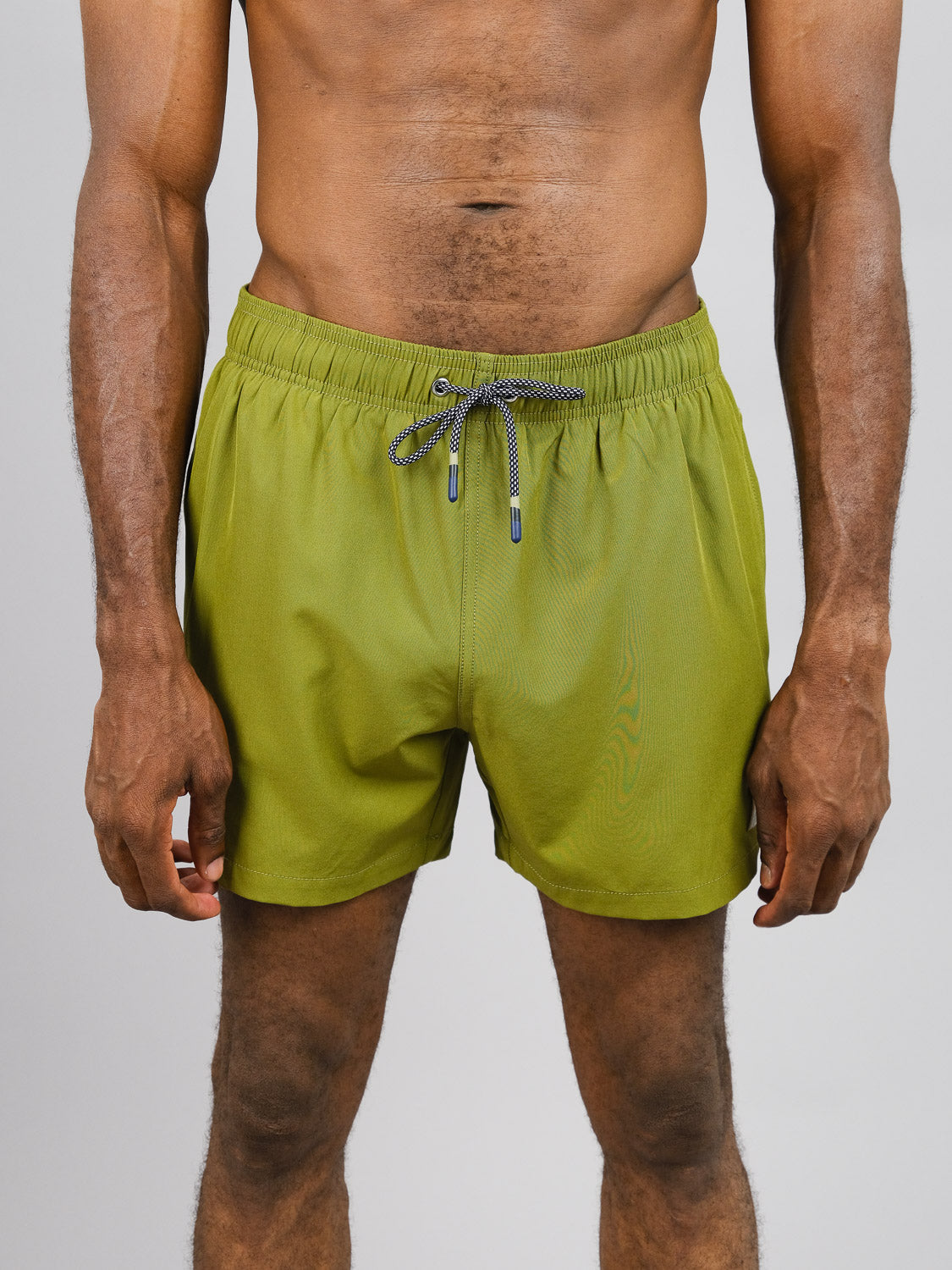 A person stands against a plain background, modeling the Oyogu Quick Dry 4 Way Stretch Swim Trunk from ID Menswear in olive green. Their hands rest by their sides, showcasing the textured fit of these shorts crafted from recycled plastic bottles.