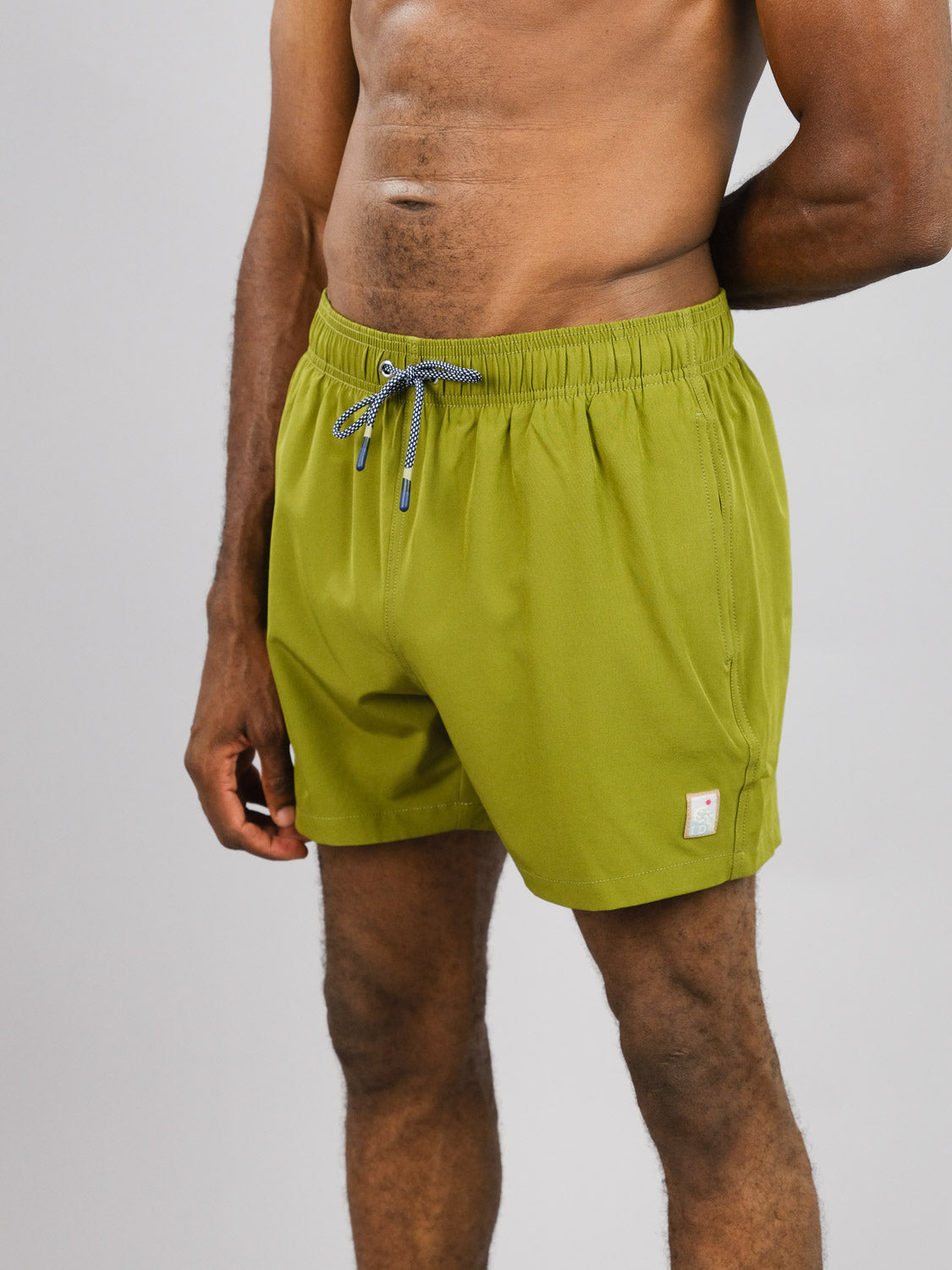 Against a plain gray background, a person wears Oyogu Quick Dry 4 Way Stretch Swim Trunk by ID Menswear, made from recycled plastic bottles. These olive green shorts feature a drawstring and emblem near the hem, showcasing their quick-dry capabilities on the lower half of the body.