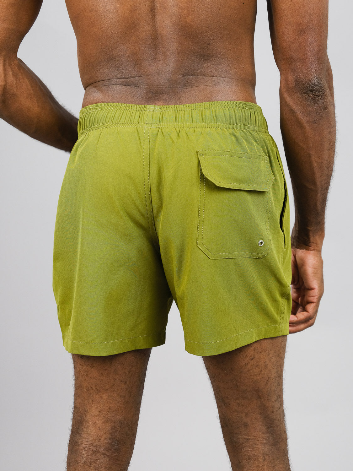 Someone wears olive green Oyogu Quick Dry 4 Way Stretch Swim Trunks by ID Menswear, crafted from recycled plastic bottles. The shorts feature an elastic waistband and a flap pocket on one side, standing out against a plain gray background.