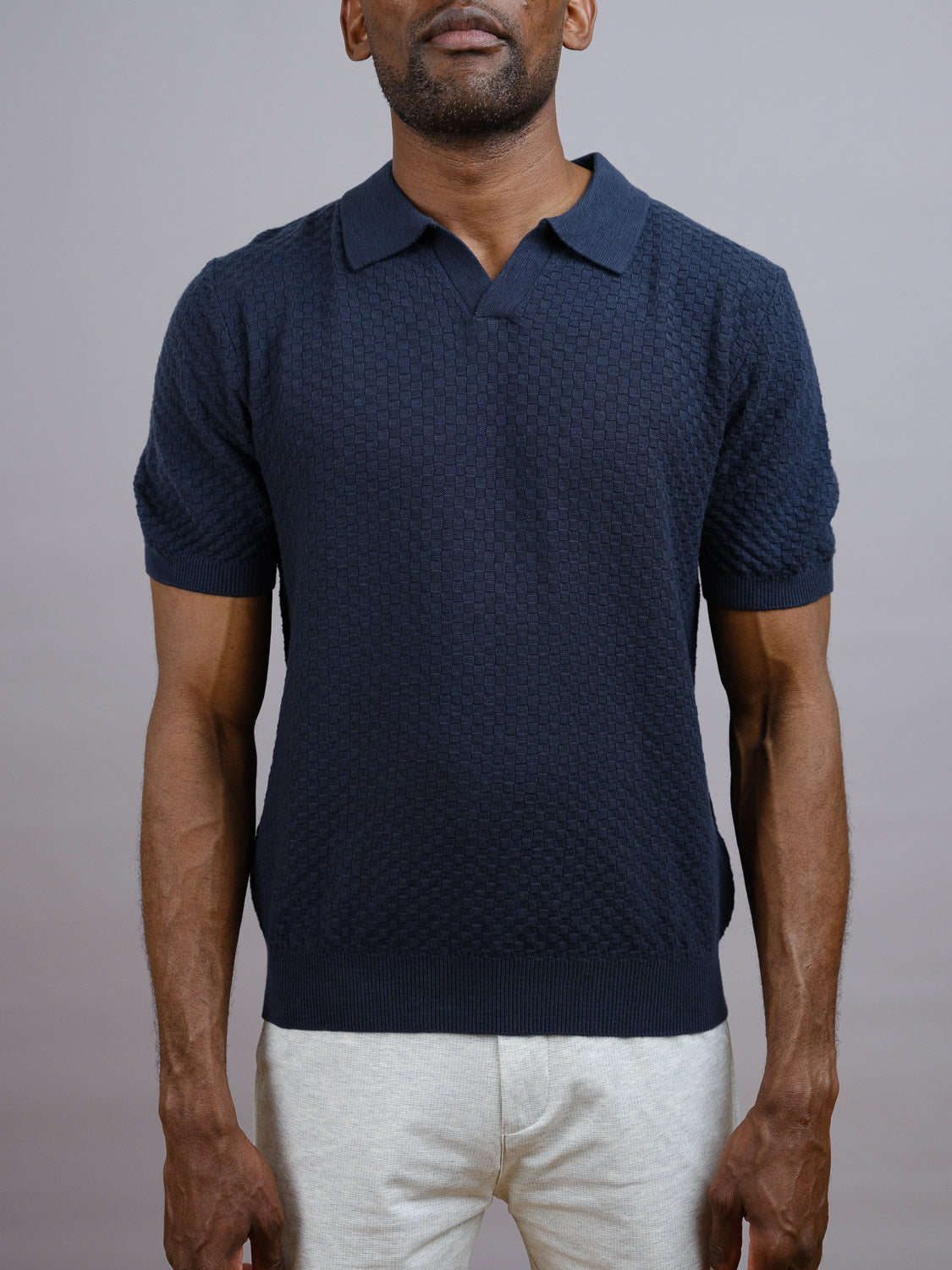A man wears the ID Menswear Cairo Knitted Cotton Polo in textured navy blue with light gray pants, set against a plain gray background. His face is partly out of frame, emphasizing the premium appeal of this casual style.