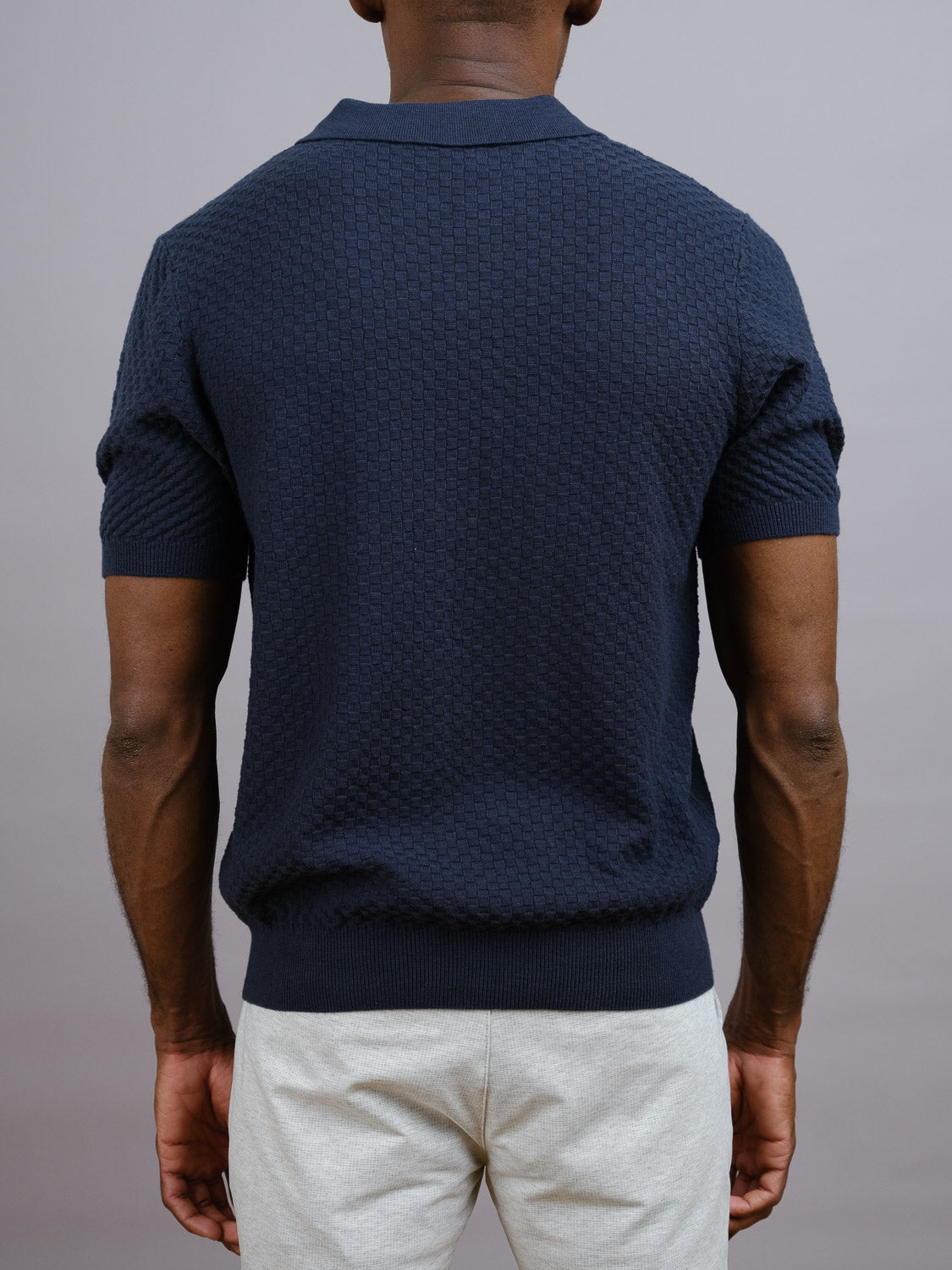A man stands with his back to the camera, wearing the Cairo Knitted Cotton Polo by ID Menswear in navy and light gray pants, set against a plain gray background for a premium casual style.
