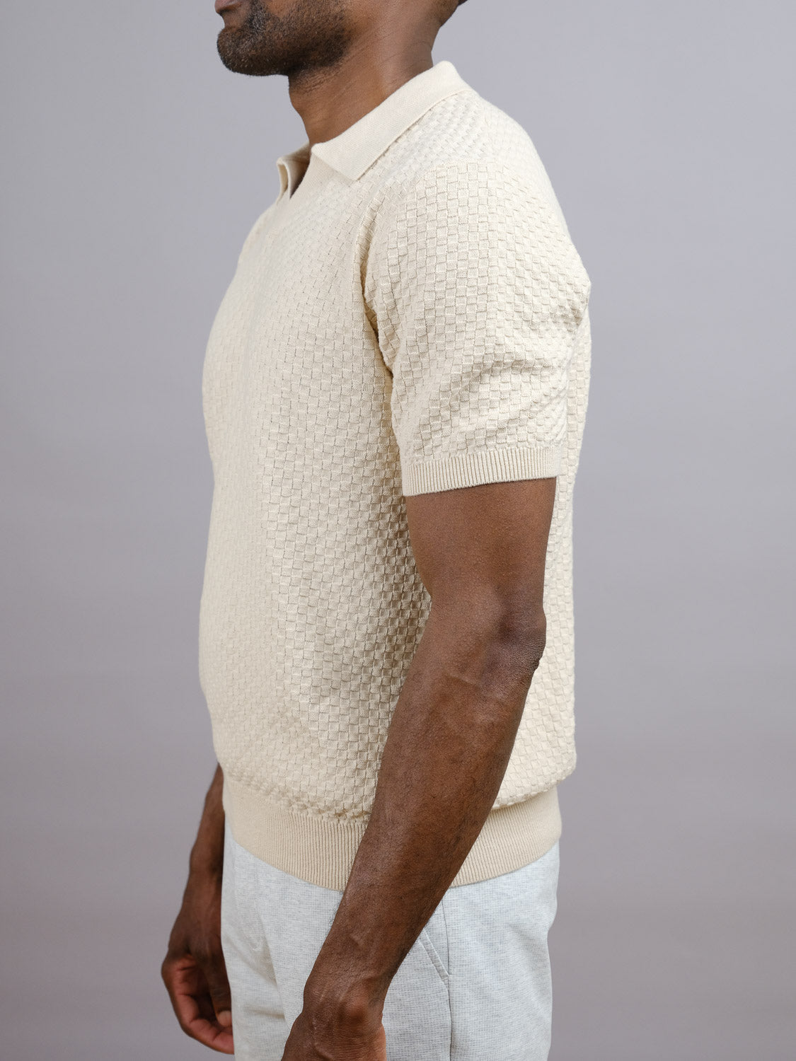 A man stands in profile wearing the Cairo Knitted Cotton Polo by ID Menswear, a premium-feel light beige cotton-knit shirt paired with light pants—an effortless take on casual style—set against a plain gray background.