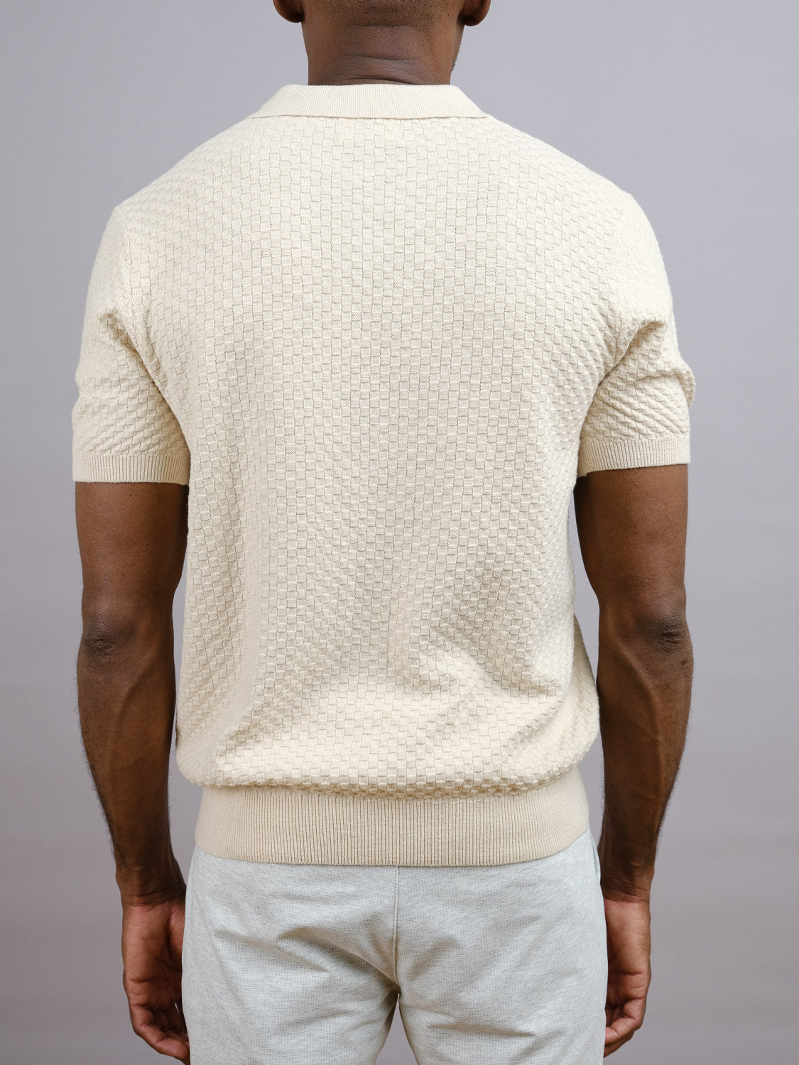 A man, back to the camera, wears the ID Menswear Cairo Knitted Cotton Polo in cream with light pants against a plain gray background, highlighting the premium comfort and modern style of this short-sleeved casual top.