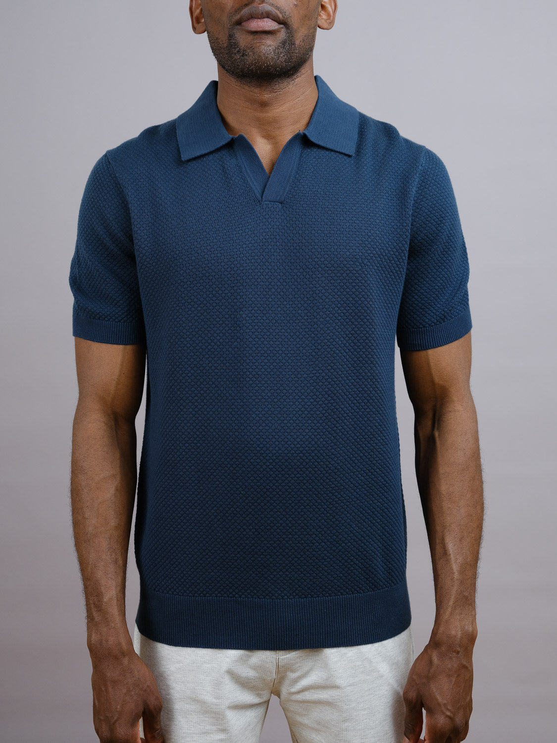 A man wears the navy Malaga Knitted Cotton Polo by ID Menswear, crafted from 100% cotton in a pineapple knit pattern, paired with light pants. He stands facing forward against a plain gray background, arms relaxed at his sides.