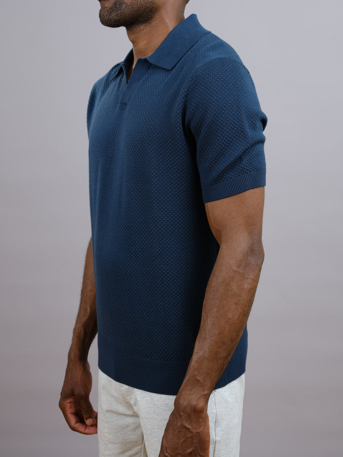 A man in the ID Menswear Malaga Knitted Cotton Polo, featuring a textured navy design and pineapple needle knit, pairs it with light pants as he stands against a plain gray background.