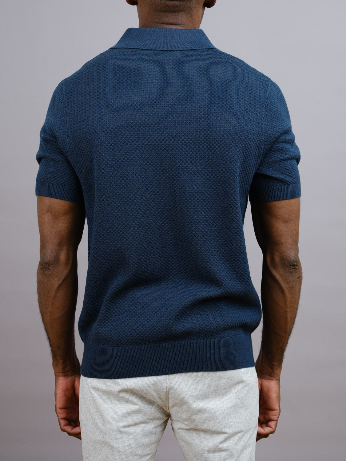 A man faces away from the camera, wearing the navy Malaga Knitted Cotton Polo by ID Menswear—made from 100% cotton with a textured pineapple needle pattern—and light pants, against a plain gray background.