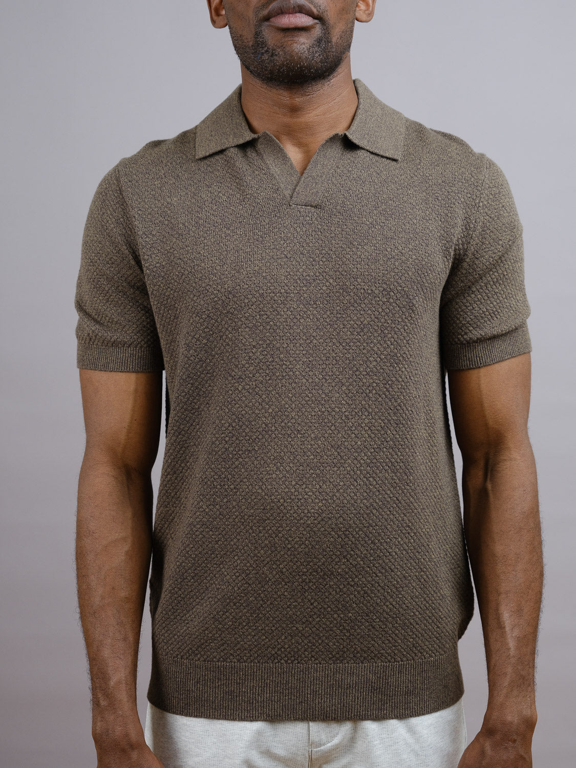 A man wears the ID Menswear Malaga Knitted Cotton Polo, a short-sleeve, textured 100% cotton shirt with a collar and open neckline, paired with light pants. He stands against a plain, light gray background.