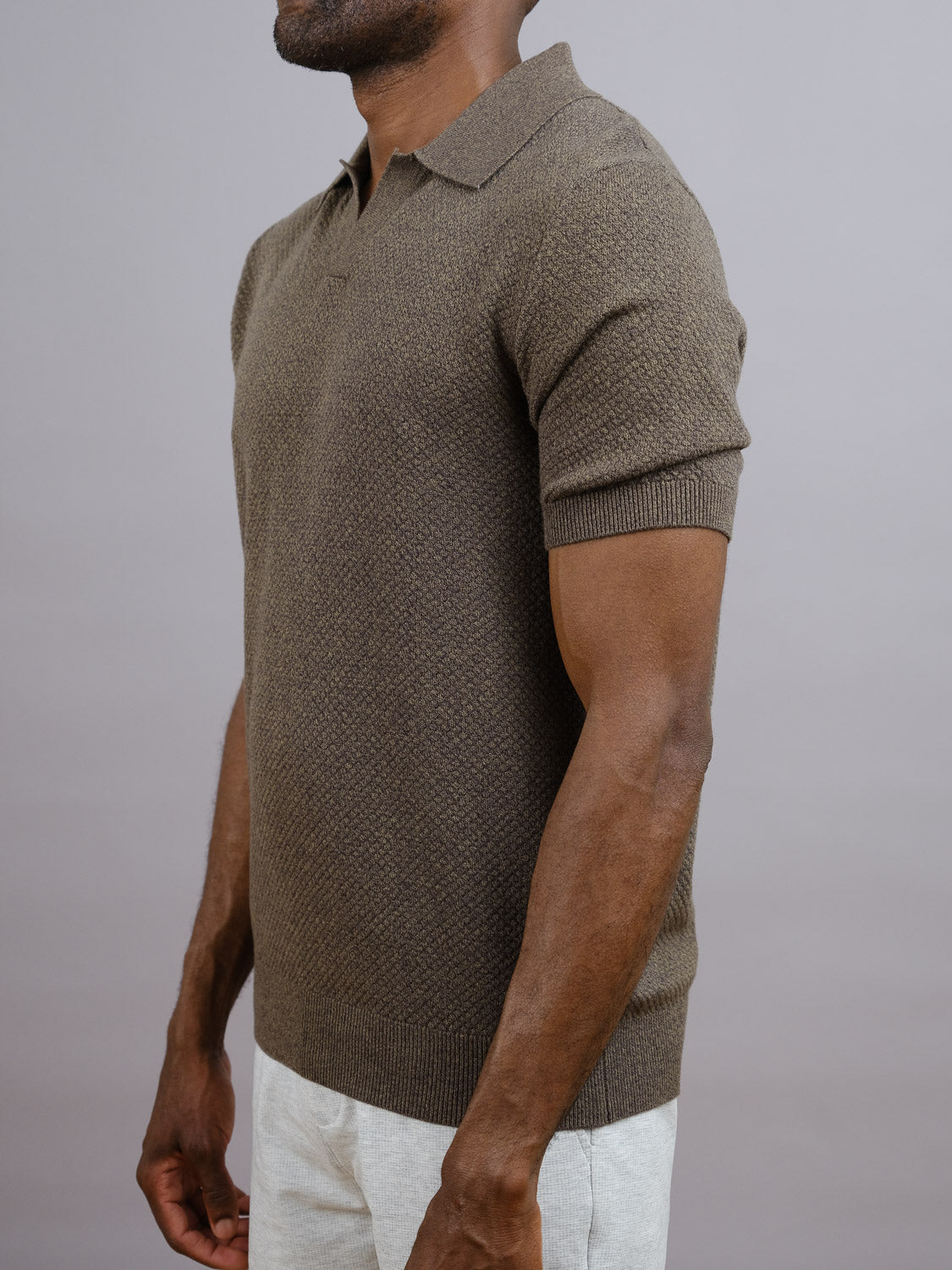 A man wears the ID Menswear Malaga Knitted Cotton Polo in brown, featuring short sleeves and textured 100% cotton fabric, paired with light pants. The photo is cropped to show only his torso and arms against a plain gray background.