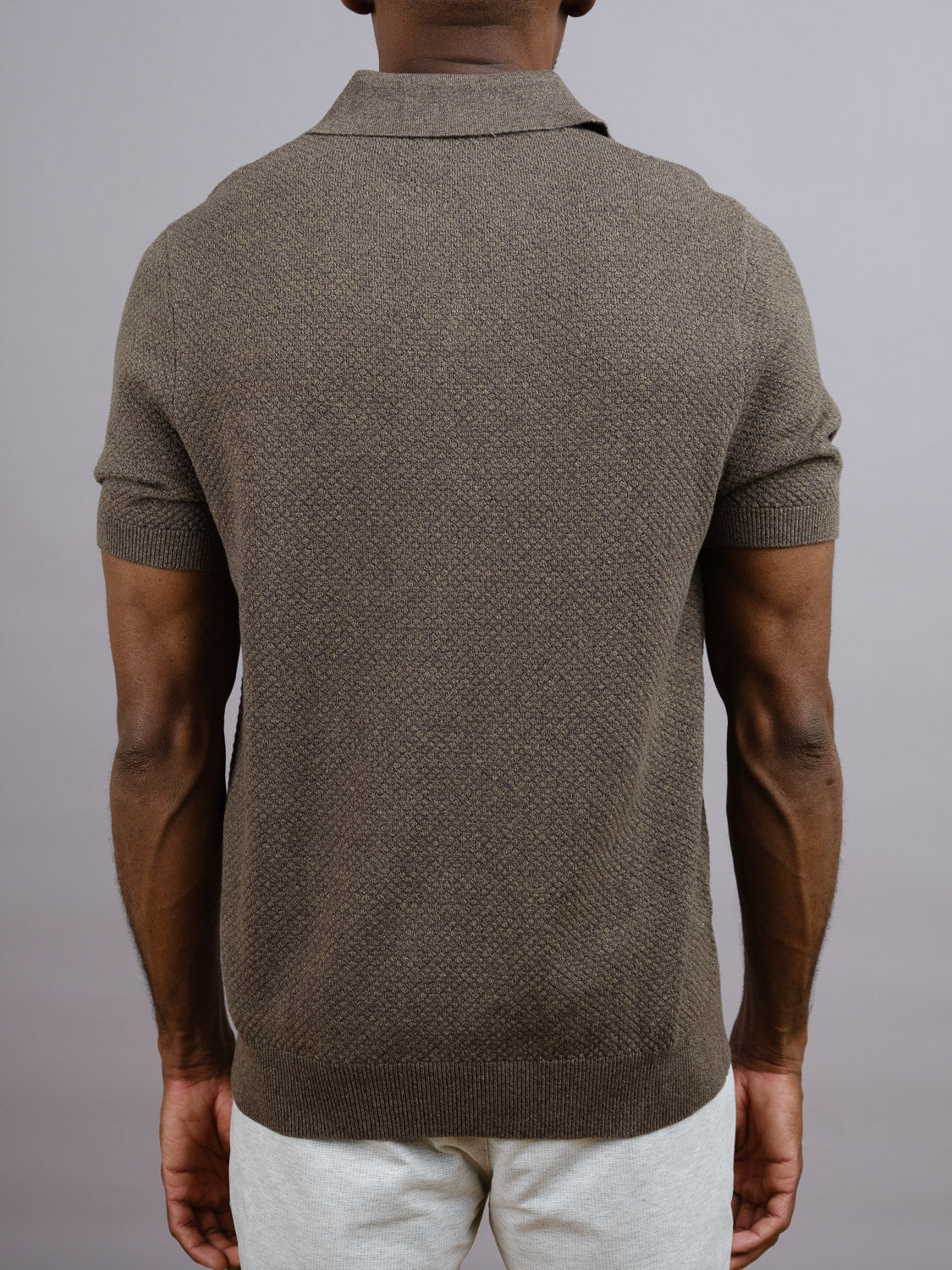 A man seen from behind wears the ID Menswear Malaga Knitted Cotton Polo, featuring a short-sleeve 100% cotton design with a pineapple needle texture, paired with light pants against a plain gray background.