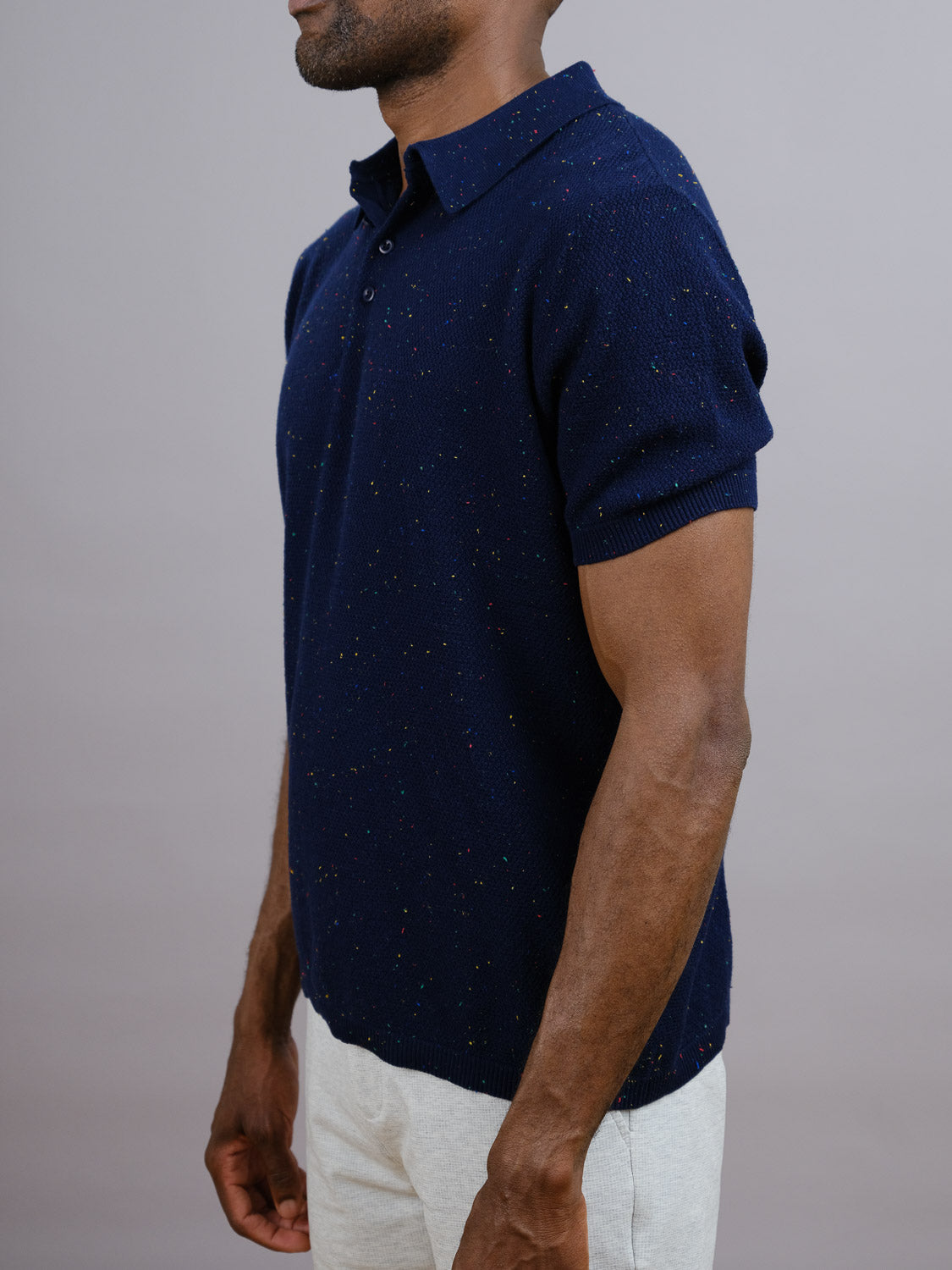 A man stands in profile against a gray background wearing the New Austin Knitted Cotton Polo by ID Menswear—a refined, short-sleeve casual style in navy speckled cotton—paired with light pants for a modern look.