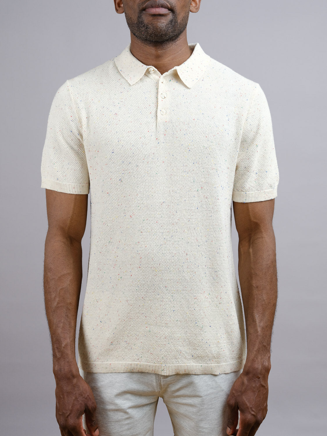 A person wears the New Austin Knitted Cotton Polo by ID Menswear in light beige, featuring short sleeves and speckled cotton, standing against a plain gray background to showcase versatile casual style.