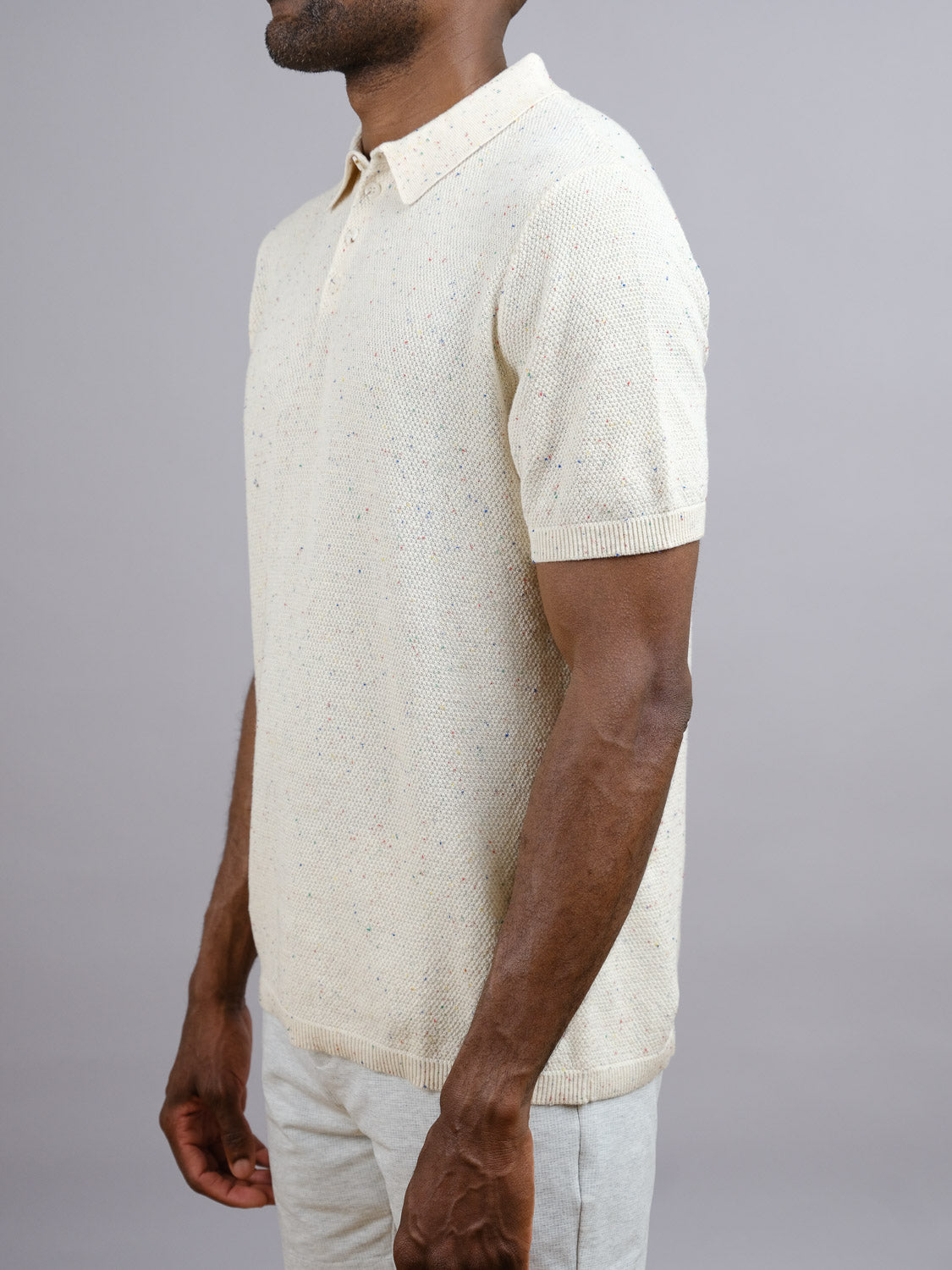 A man wears the New Austin Knitted Cotton Polo by ID Menswear in light beige with short sleeves, made from speckled cotton. He stands against a plain gray background, head turned slightly, highlighting this timeless casual piece.