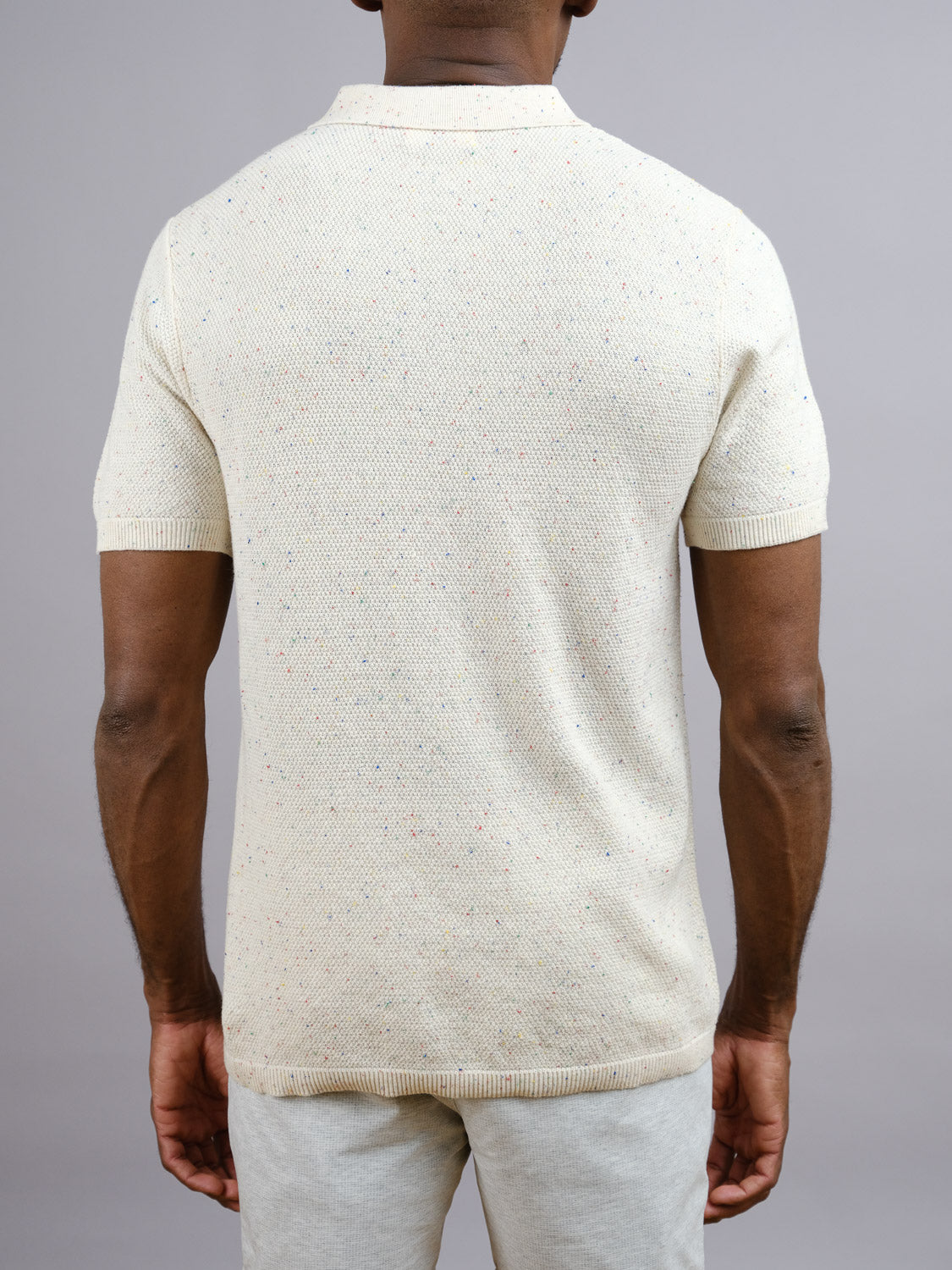 A person, back to the camera, wears a short-sleeved cream New Austin Knitted Cotton Polo by ID Menswear and light pants—an easy casual choice—against a plain gray background.