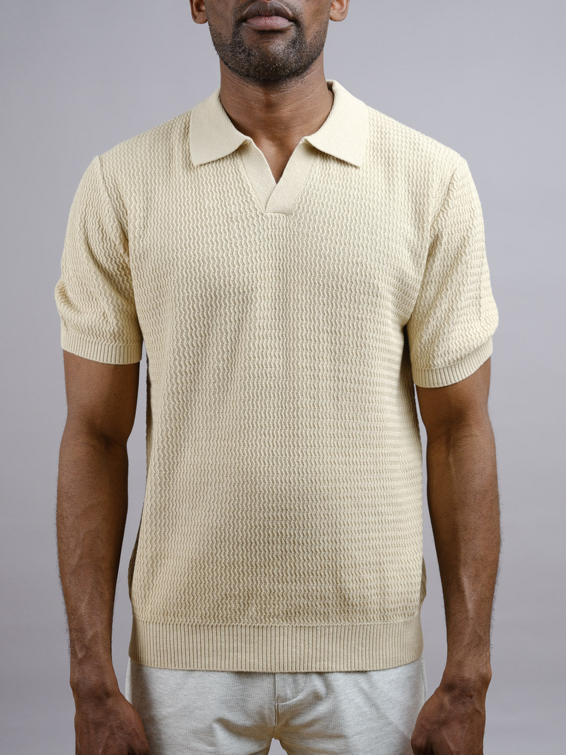 A man models the Mecca Linen Blend Knit Polo by ID Menswear with light-colored pants against a plain gray background; his face is partially out of frame, arms resting casually at his sides.
