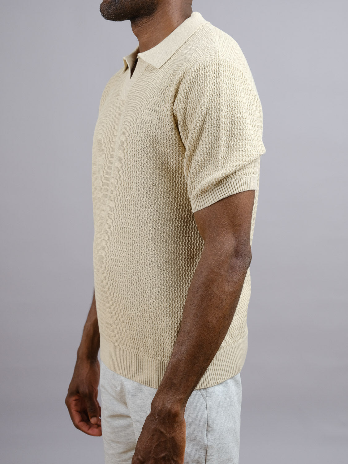A man in the Mecca Linen Blend Knit Polo by ID Menswear, featuring a lightweight breathable fabric with a structured wave knit, pairs it with light-colored pants and stands sideways against a plain gray background.