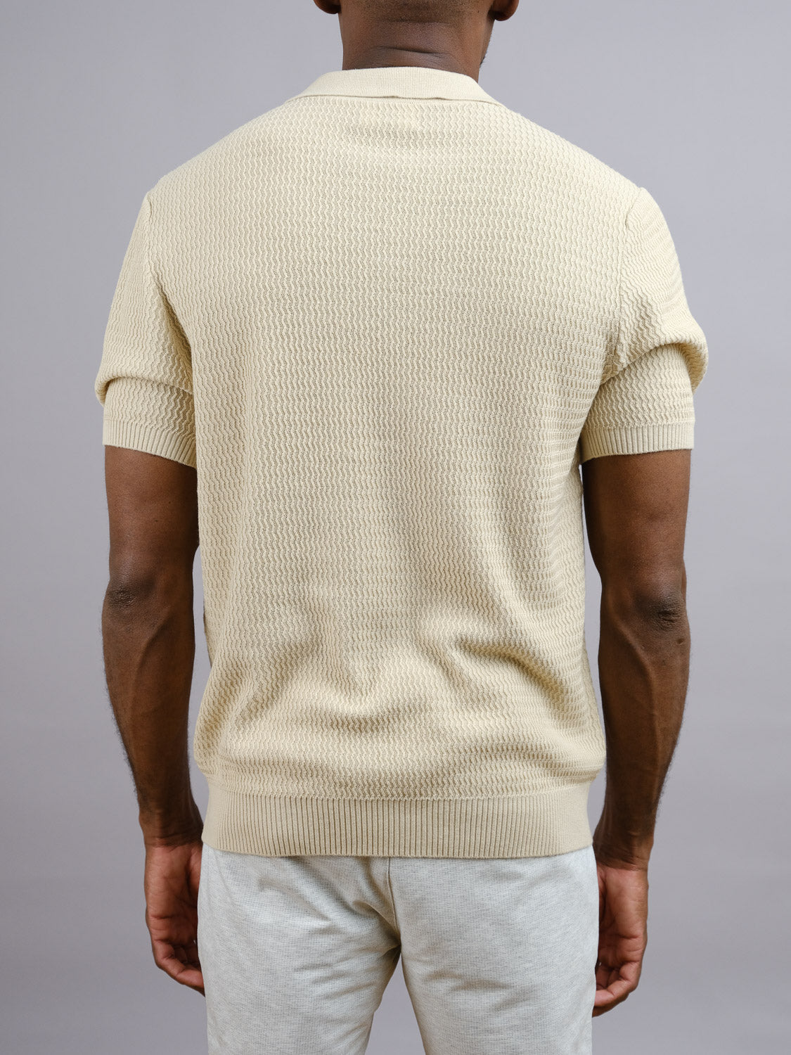 A man facing away from the camera wears the Mecca Linen Blend Knit Polo by ID Menswear and light-colored pants against a plain gray background.
