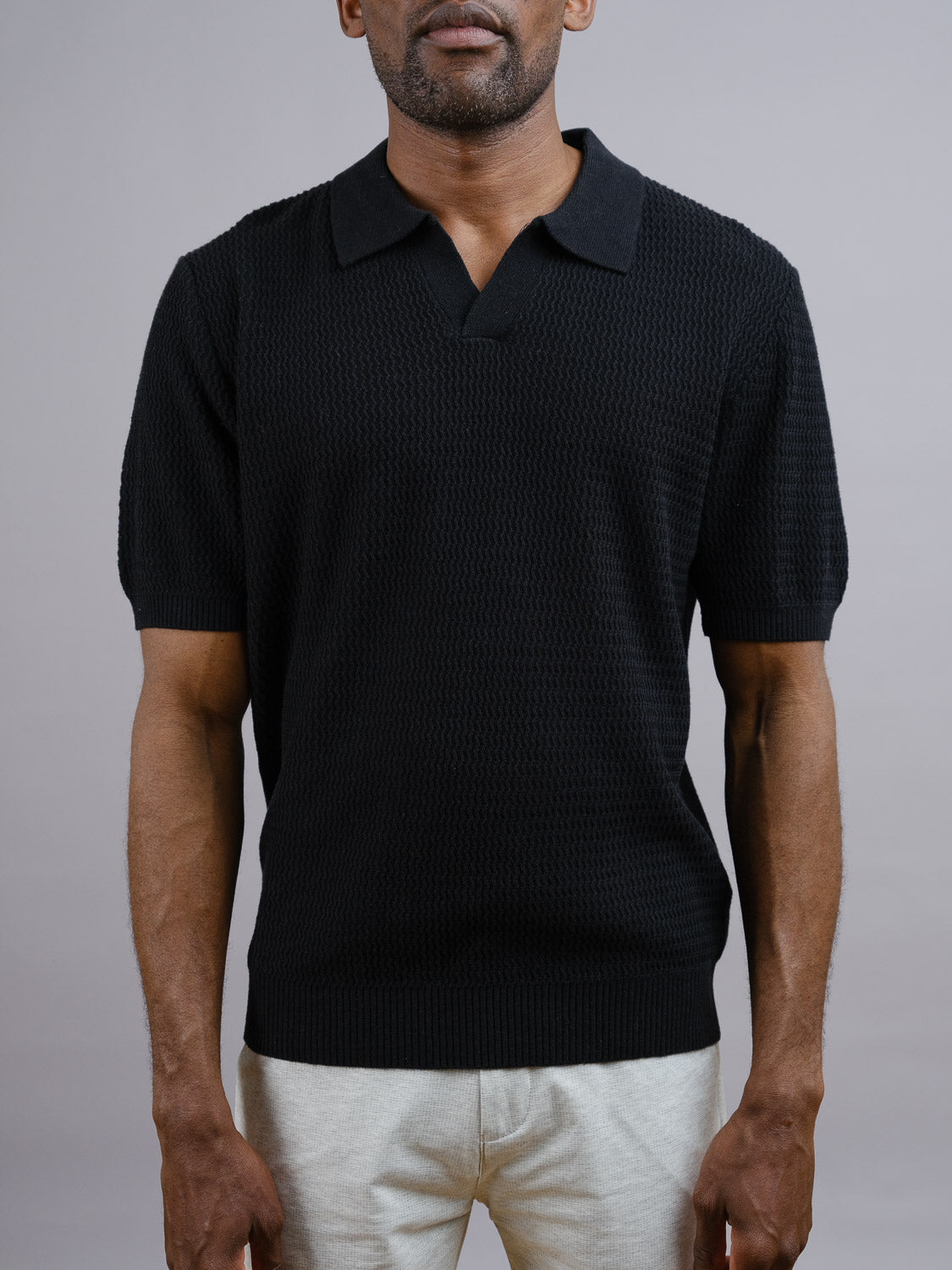 A man wears the black Mecca Linen Blend Knit Polo by ID Menswear with light-colored pants, standing against a plain gray background. His head is partially out of frame.