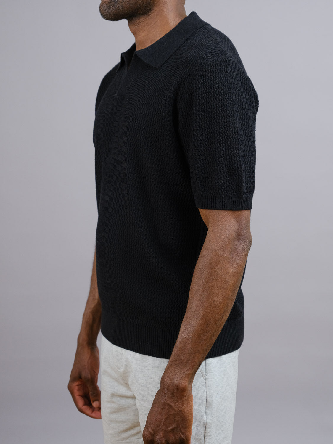 A man models the ID Menswear Mecca Linen Blend Knit Polo with light pants, standing sideways against a gray background. The view is from shoulders to mid-thigh, with his head turned slightly.