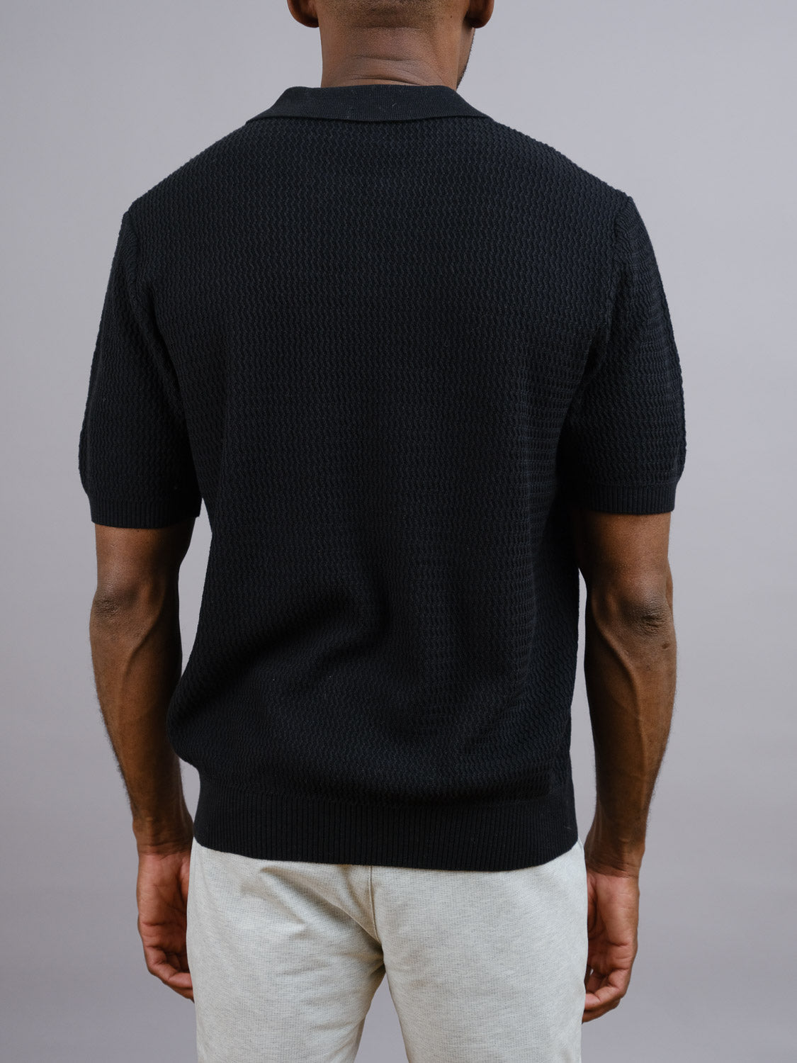 A person wearing an ID Menswear Mecca Linen Blend Knit Polo in black and light gray pants stands facing away from the camera against a plain gray background.