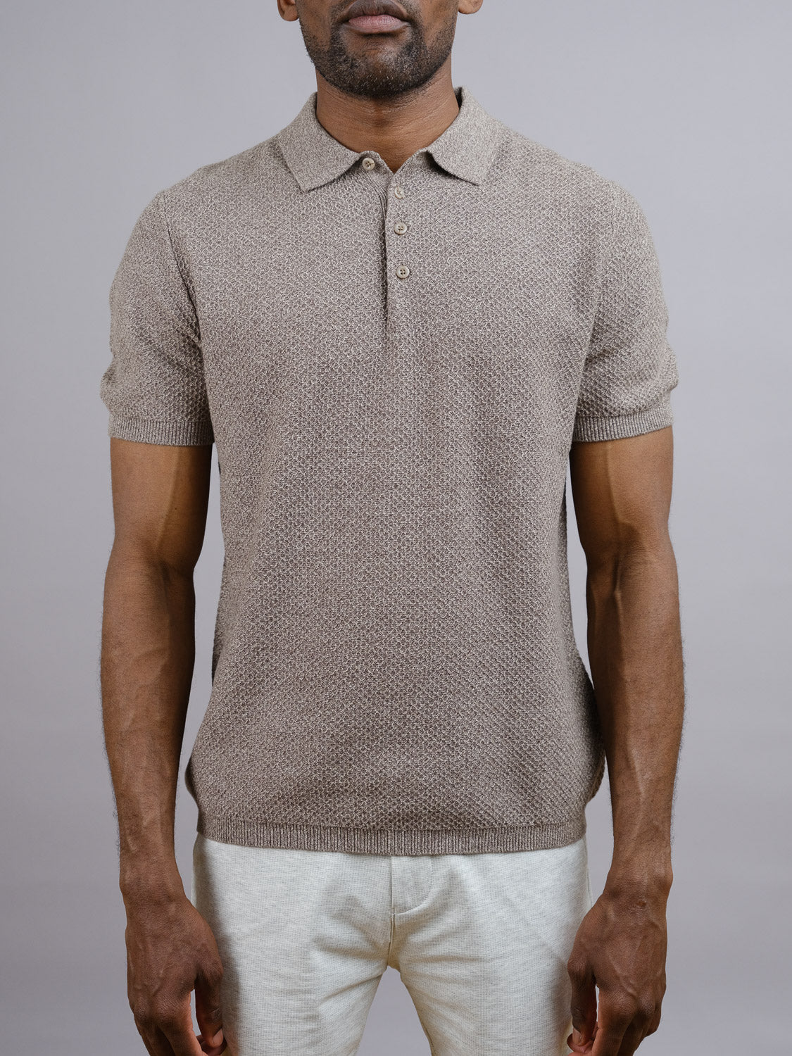 A man models the Tucson Knitted Cotton Polo by ID Menswear in textured light brown with light pants, standing against a plain gray background. His face is partially out of frame, highlighting this versatile casual piece.