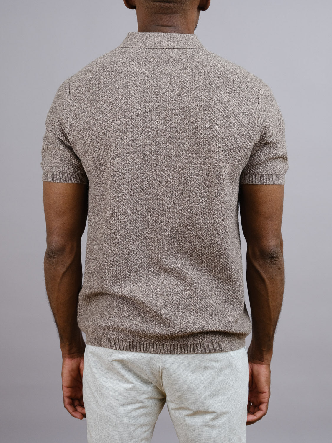 A man faces away, wearing the Tucson Knitted Cotton Polo by ID Menswear in textured light brown with light pants—exemplifying casual, breathable style against a plain gray background.