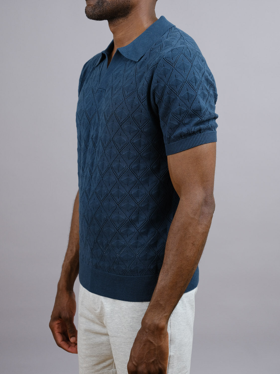 A man models the Agra Knitted Cotton Polo by ID Menswear in navy blue with light pants, standing against a plain gray background. This short-sleeved polo offers a breathable fit ideal for casual wear. His face is not shown.
