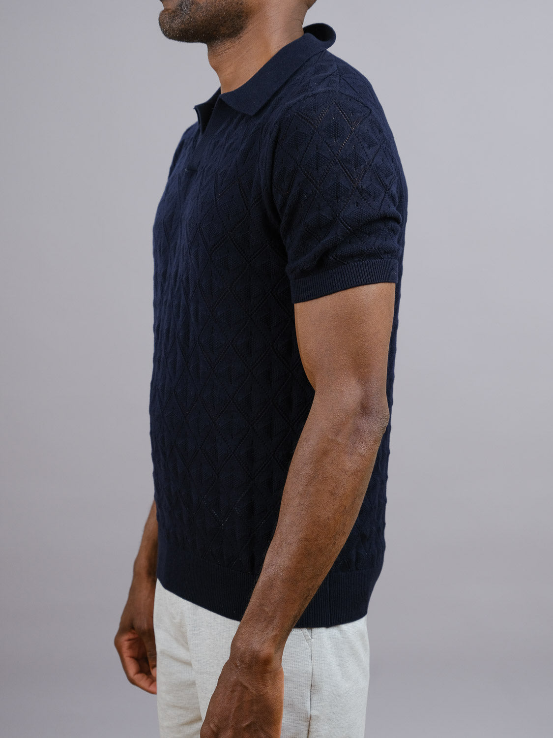 A man stands sideways against a gray backdrop, wearing the Agra Knitted Cotton Polo by ID Menswear—a short-sleeved, dark navy cotton polo with a breathable fit—paired with light pants for an understated casual look.