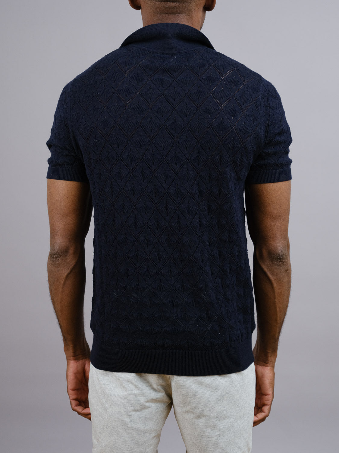A person is seen from the back wearing ID Menswear’s Agra Knitted Cotton Polo in dark blue with light pants, set against a plain gray background for a comfortable, casual look.