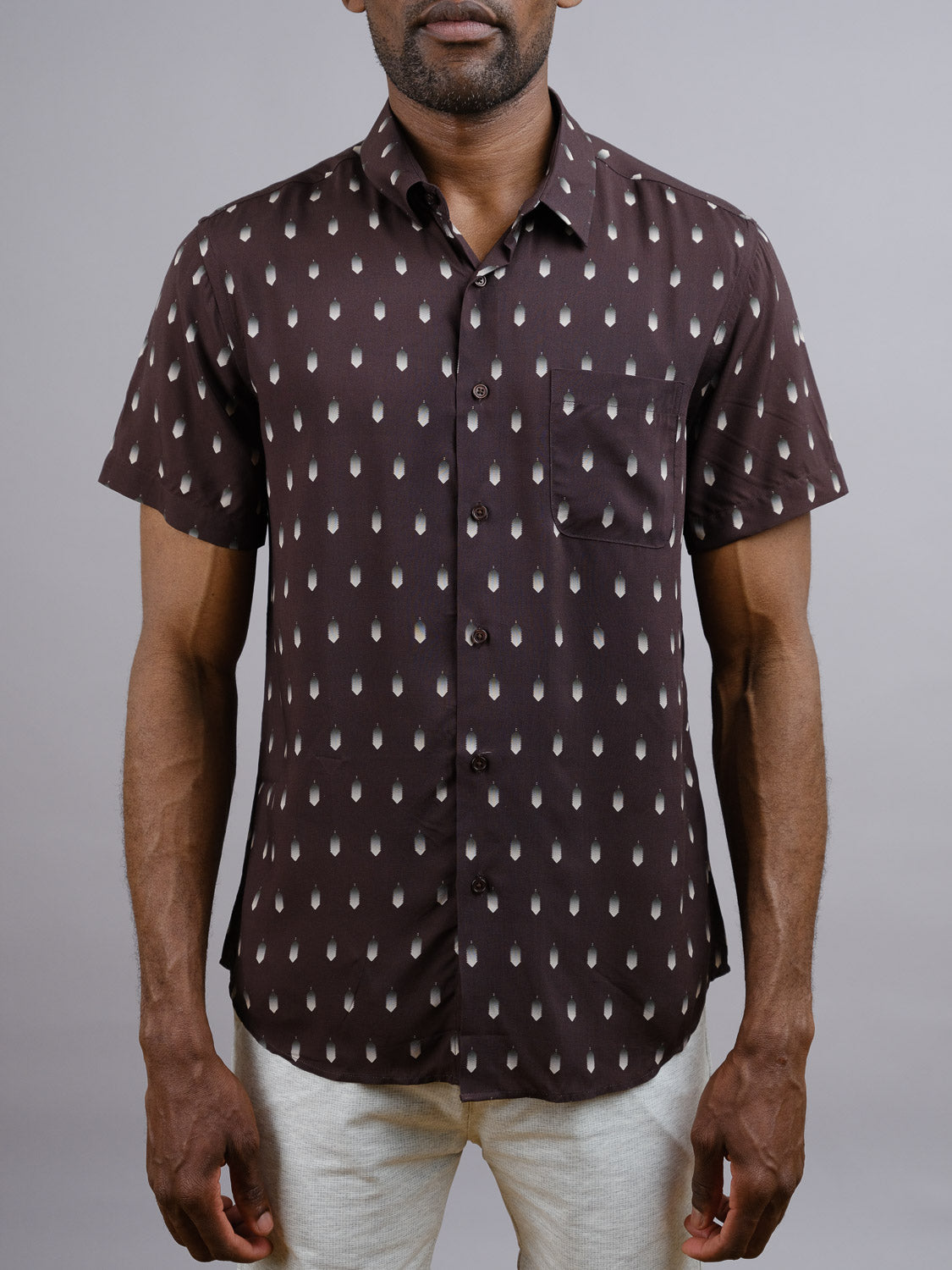 A man wears the ID Menswear Drops Printed Rayon Shirt, a short-sleeved, button-up dark brown shirt with white teardrop pattern and chest pocket, perfect for warm weather, standing against a plain light background.