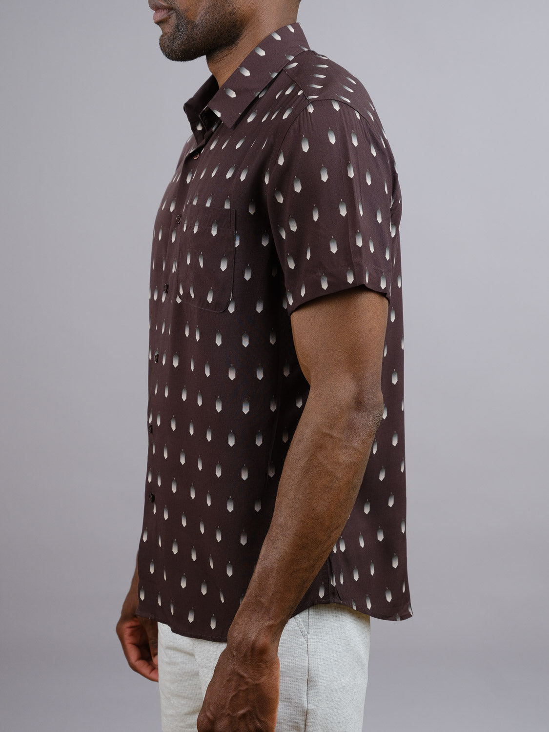 A man stands sideways against a gray background wearing the ID Menswear Drops Printed Rayon Shirt, a short-sleeve dark brown shirt with a light teardrop pattern and front pocket, paired with light-colored pants—ideal for warm weather.