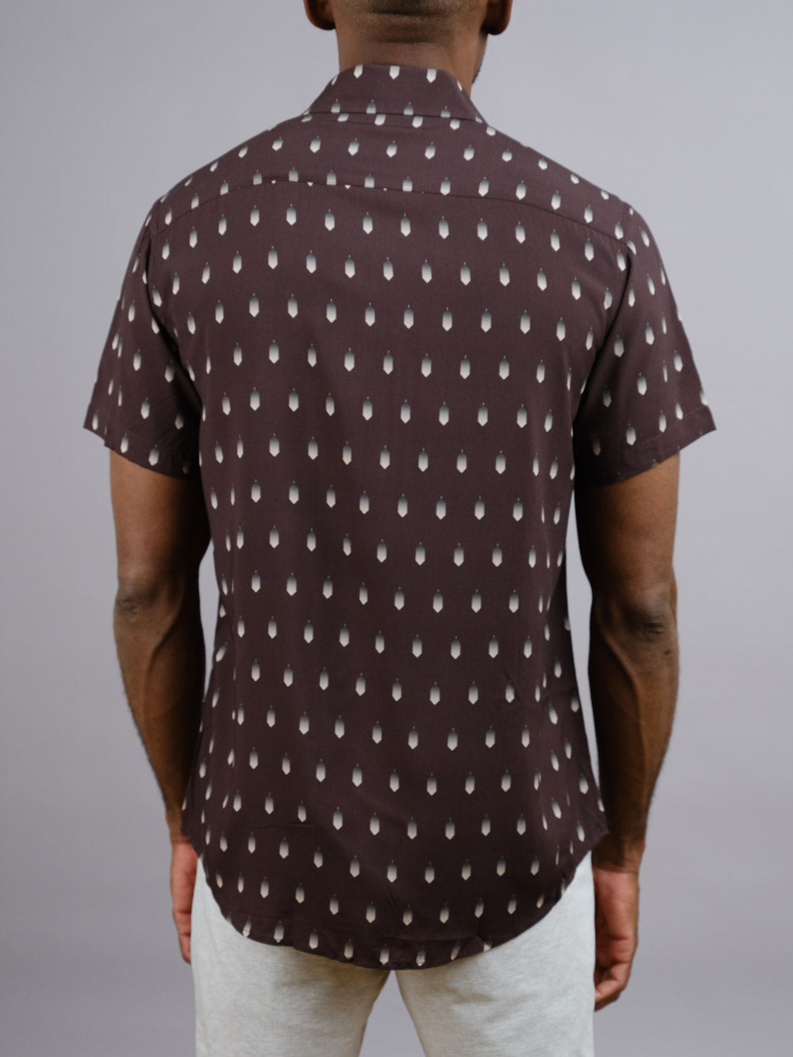 A person is seen from behind wearing the ID Menswear Drops Printed Rayon Shirt, a short-sleeve dark brown rayon shirt with a light teardrop pattern, and light grey pants, standing against a plain grey background.