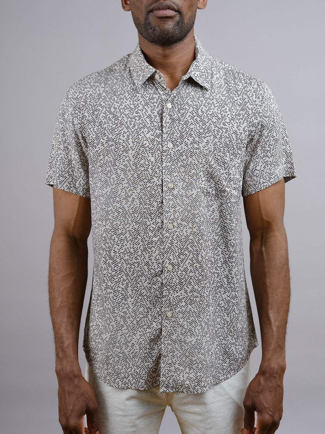 A man wears the ID Menswear Kikaga Geometric Print Shirt—short-sleeved with a black and white abstract design—untucked with light pants against a plain gray background for a relaxed, warm-weather look.