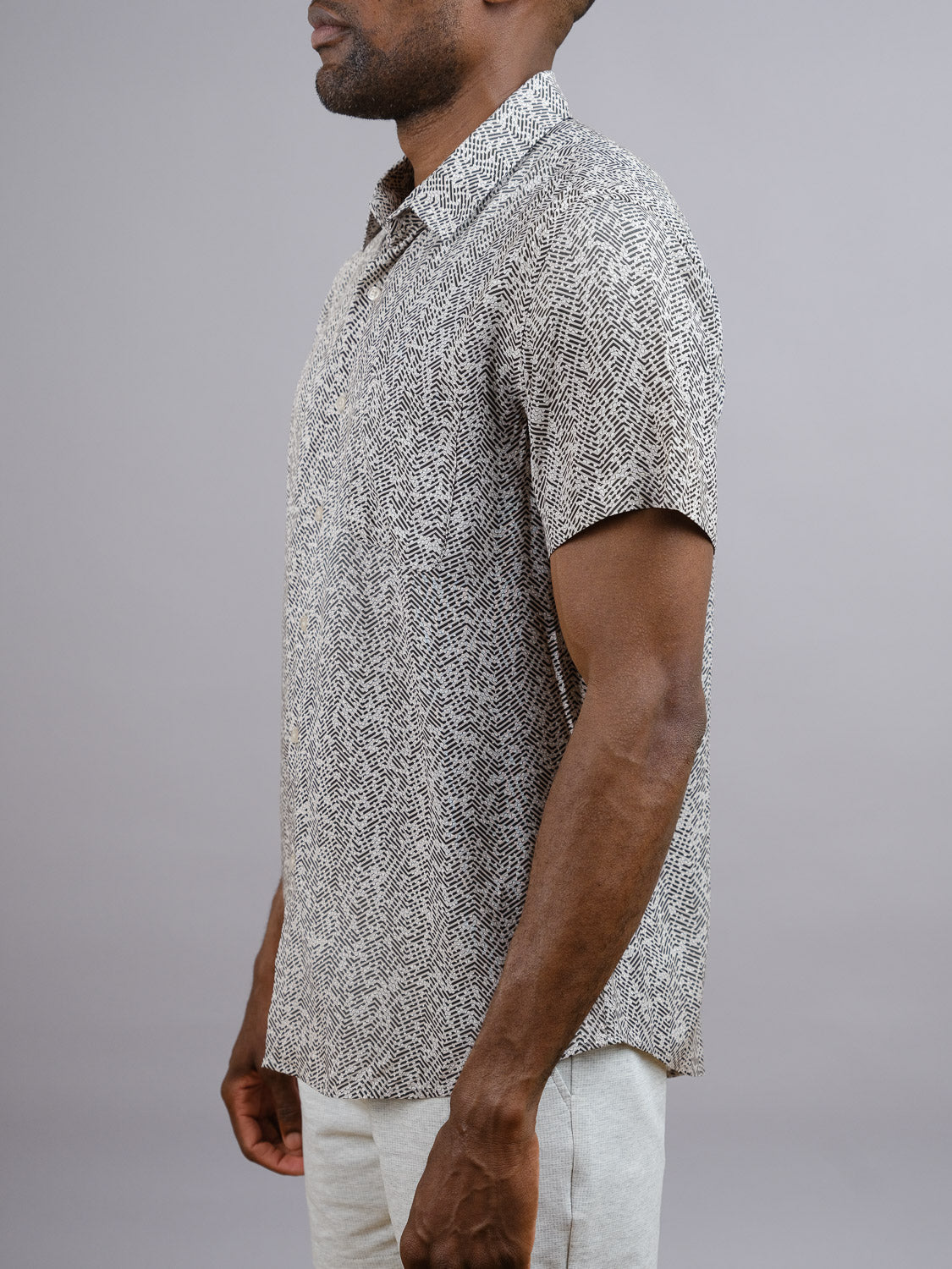 A man stands sideways in the ID Menswear Kikaga Geometric Print Shirt and light-colored pants against a plain grey background. His face is partially out of frame—a cool, vibrant look perfect for warm weather.