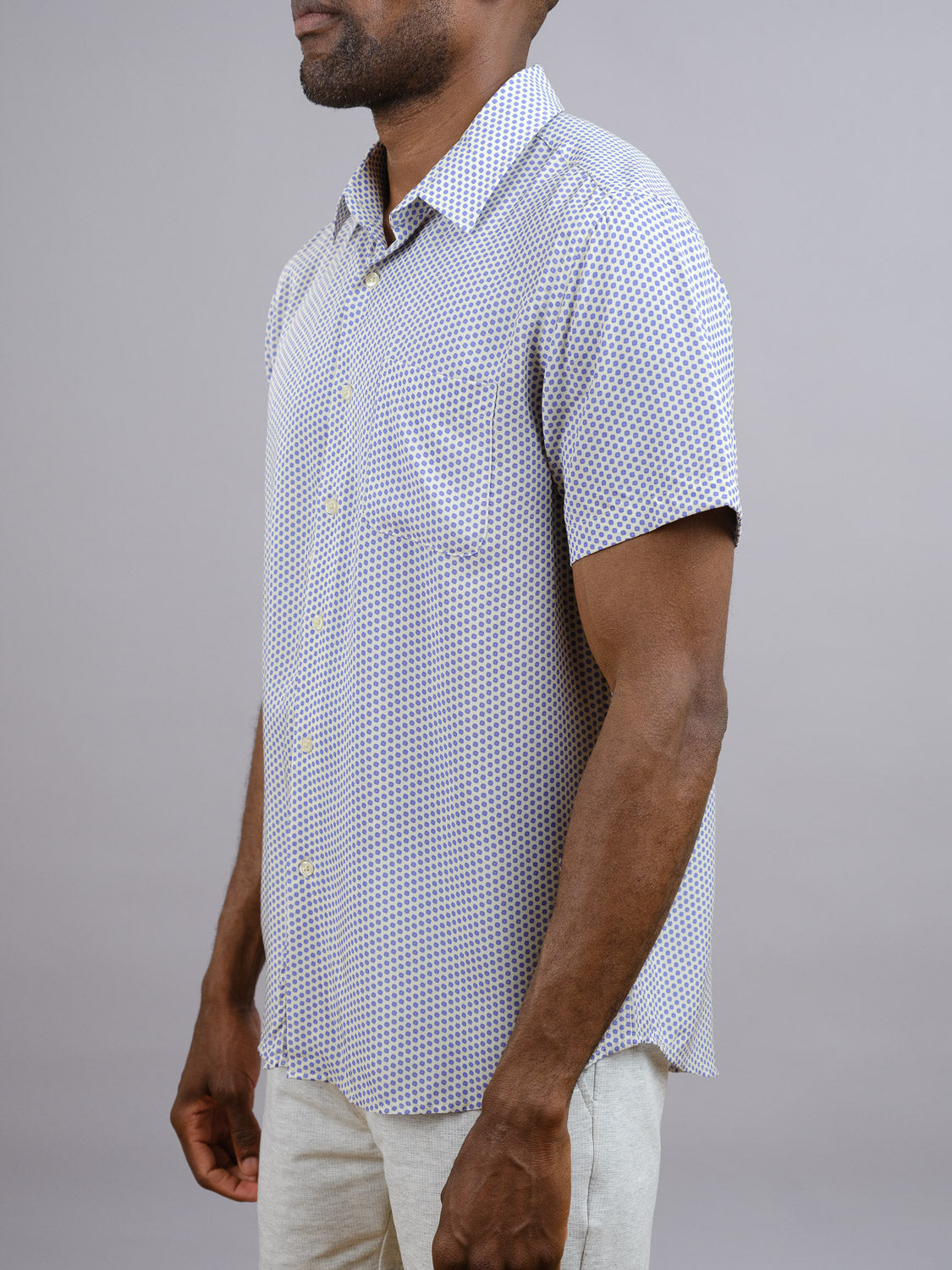 A man stands sideways against a plain gray background, wearing the Shibori Japanese Print Shirt by ID Menswear—a short-sleeve rayon shirt with a subtle geometric pattern—paired with light-colored pants, perfect for warm weather.