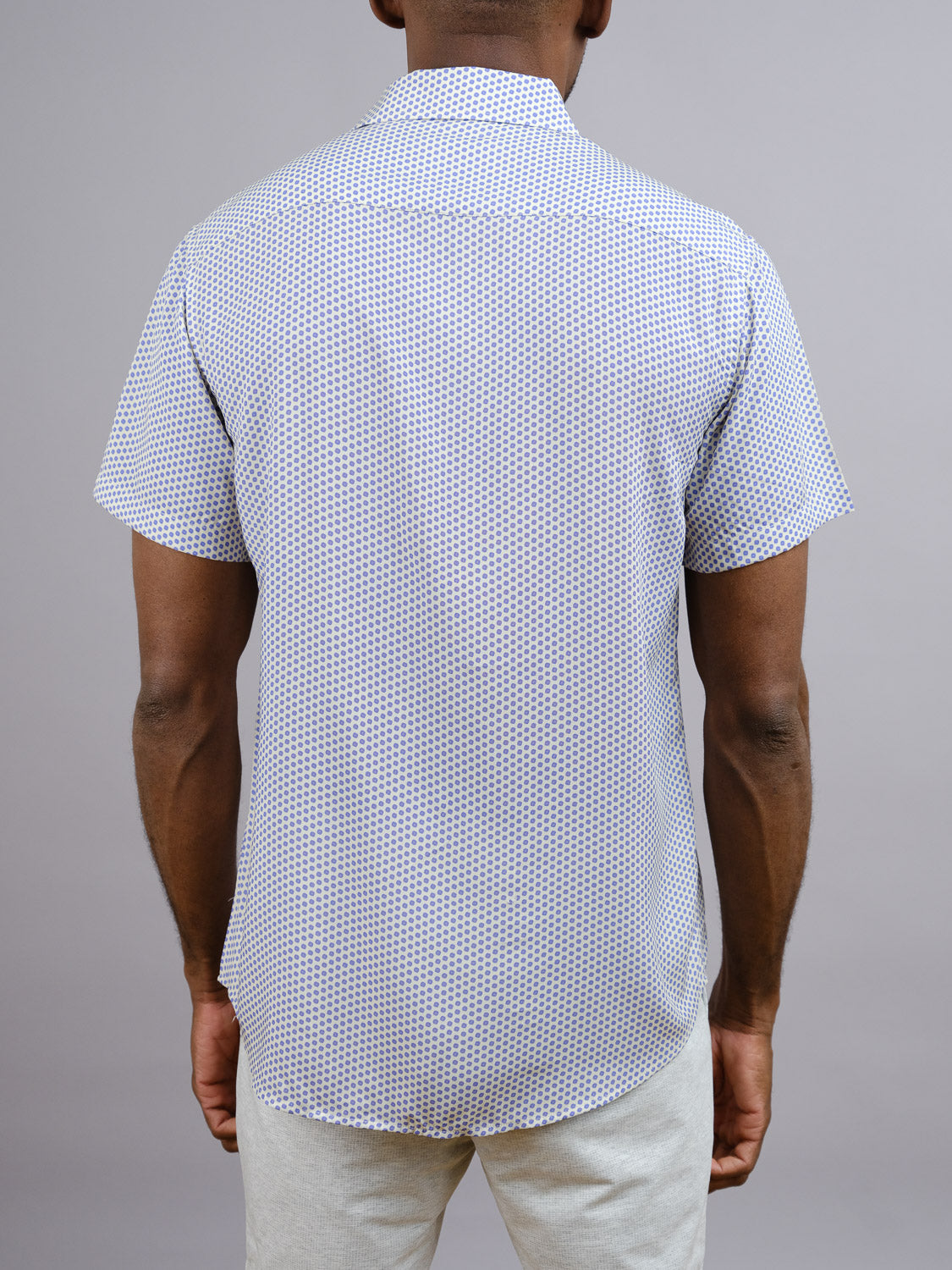 A man stands with his back to the camera, wearing the Shibori Japanese Print Shirt by ID Menswear—a short-sleeve rayon shirt with a subtle geometric pattern—paired with light pants, against a plain gray background.