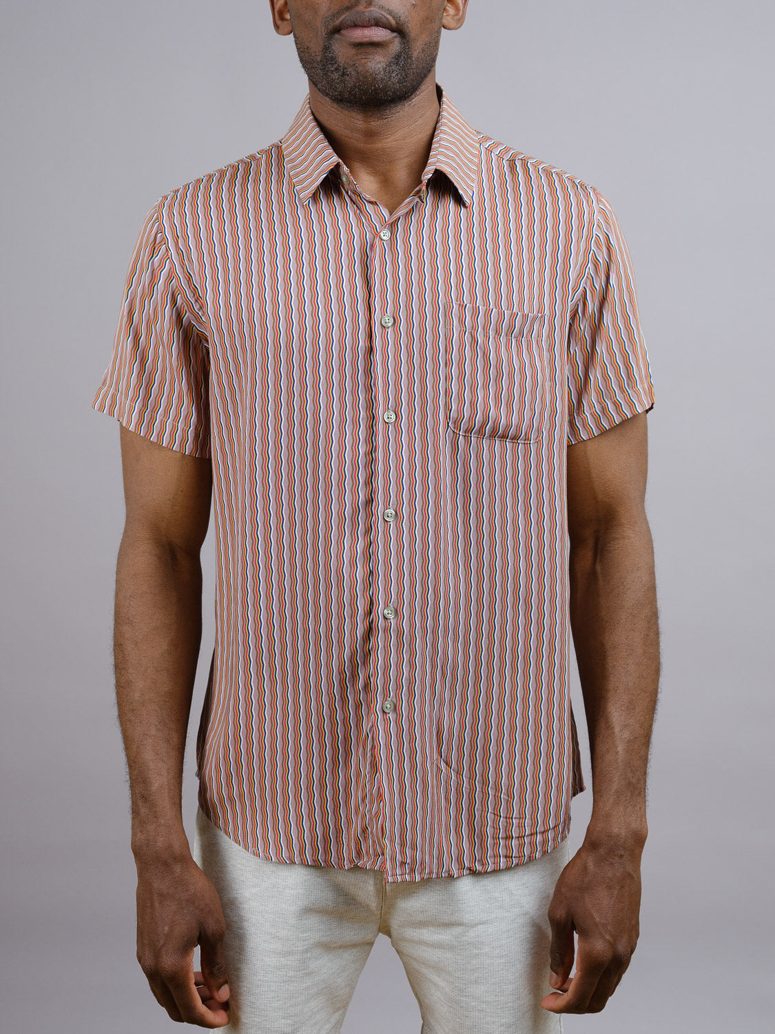 A man stands against a plain gray background wearing the Jigu Geometric Printed Shirt by ID Menswear, a short-sleeve rayon style with red, white, and blue vertical stripes—perfect for warm weather. His hands rest relaxed by his sides.