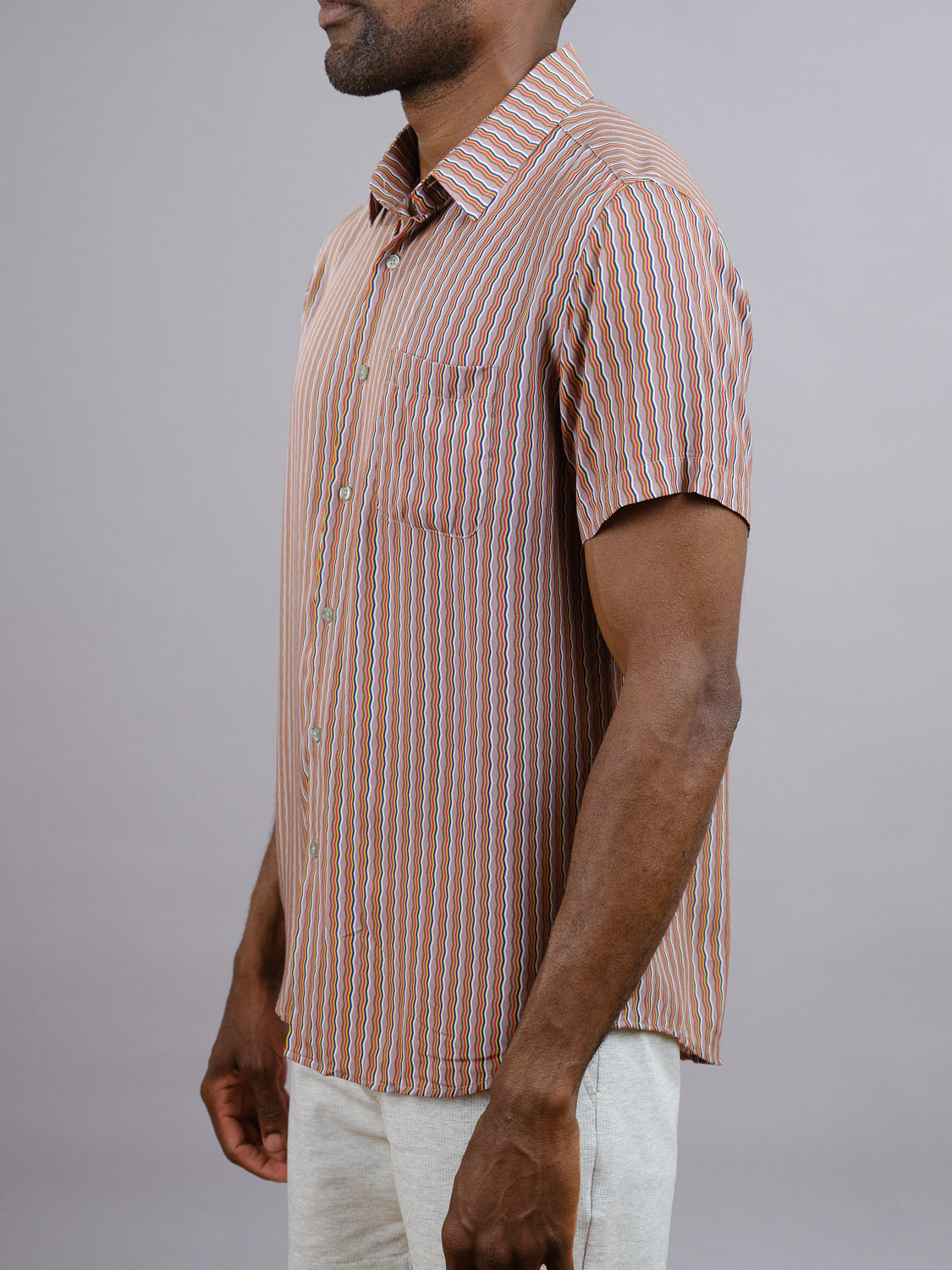 A man wearing the ID Menswear Jigu Geometric Printed Shirt—featuring short sleeves and vertical stripes in brown, white, and blue—stands sideways against a plain grey background, showcasing an ideal warm-weather look.
