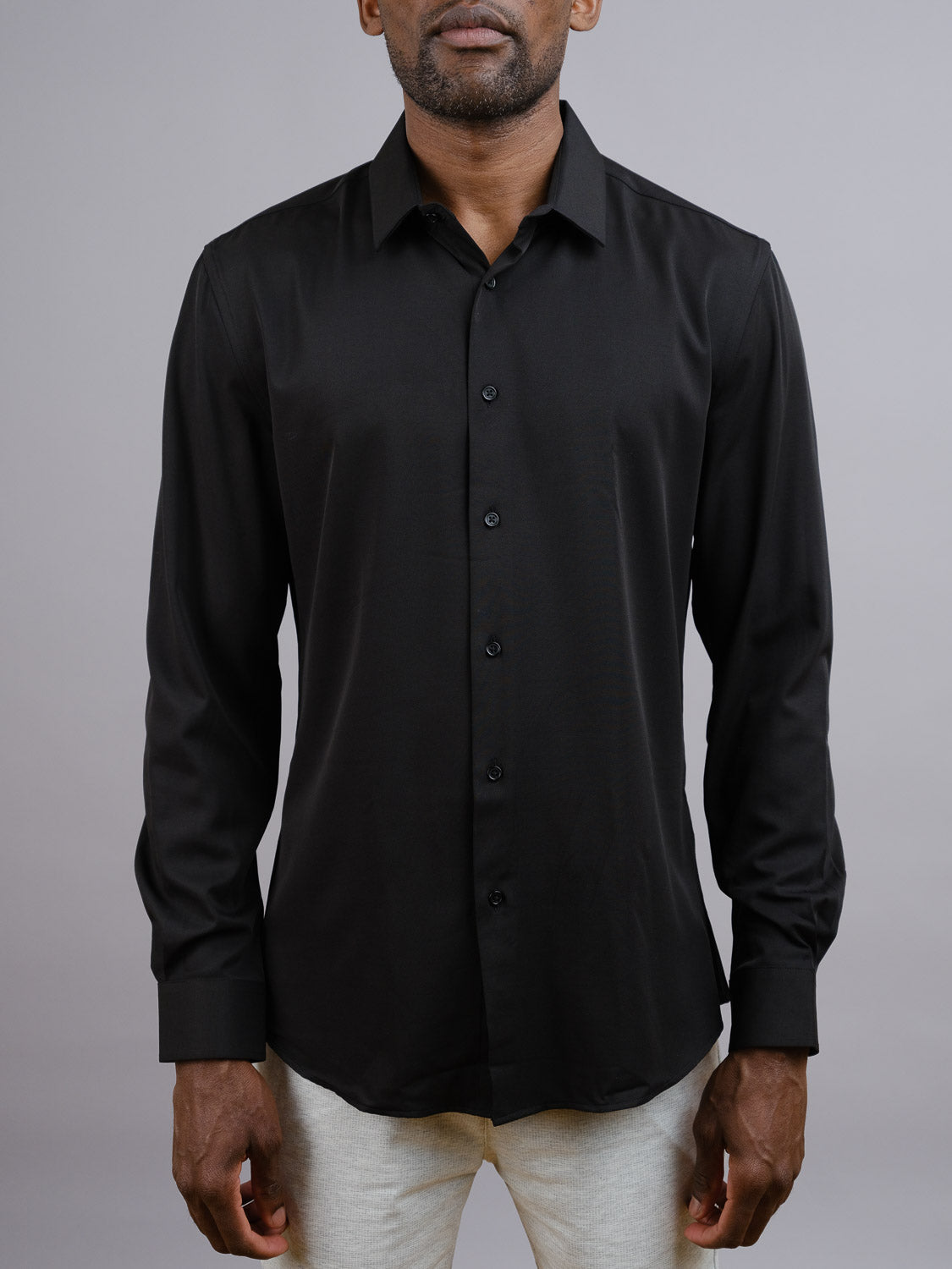 A man stands against a plain gray background wearing the ID Menswear Alex 4-Way Stretch Slim Dress Shirt in black with light-colored pants. His face is partially out of frame.