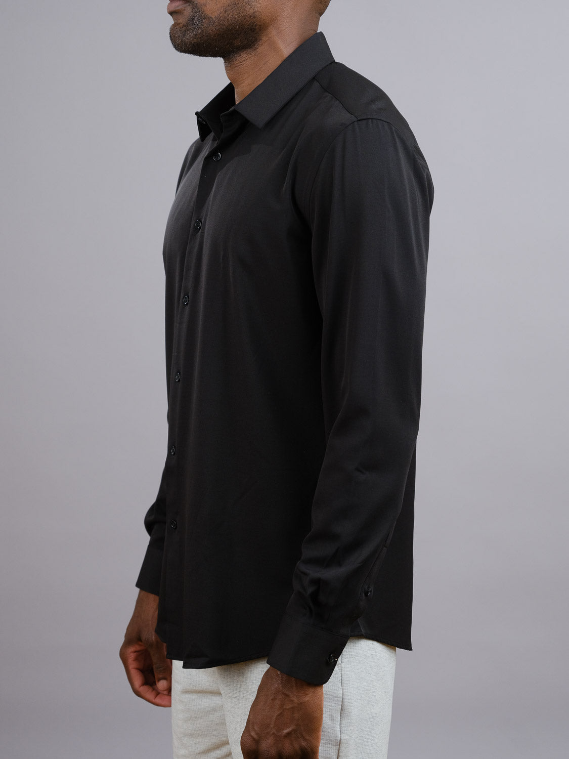 A man, shown from the side with his face partially visible, wears the ID Menswear Alex 4-Way Stretch Slim Dress Shirt in black along with light-colored pants, standing against a plain gray background.