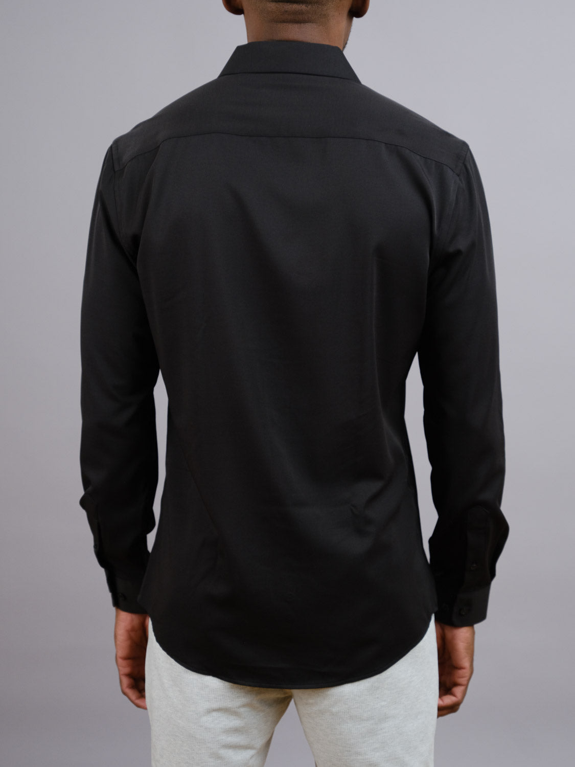 A person is shown from the back wearing the ID Menswear Alex 4-Way Stretch Slim Dress Shirt in black, paired with light-colored pants, and standing against a plain gray background.