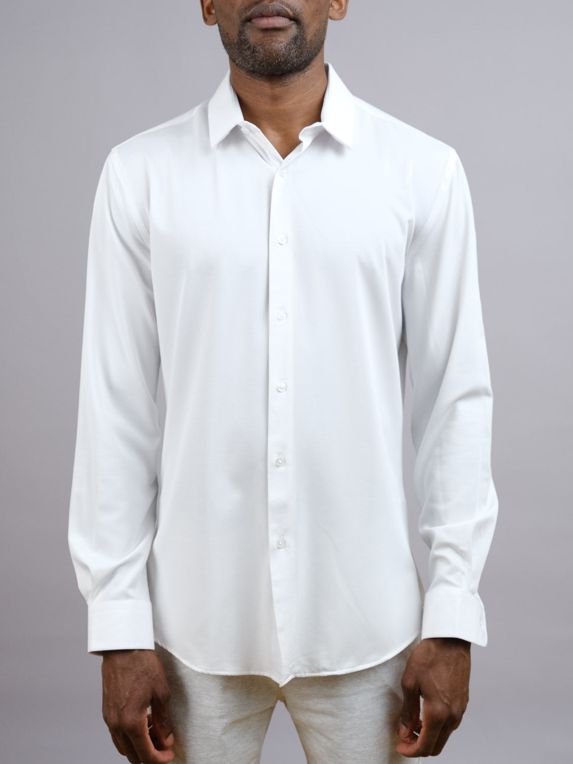A man models the ID Menswear Alex 4-Way Stretch Slim Dress Shirt in white, paired with light pants and standing against a plain gray background. His face is partially visible, arms relaxed by his sides.