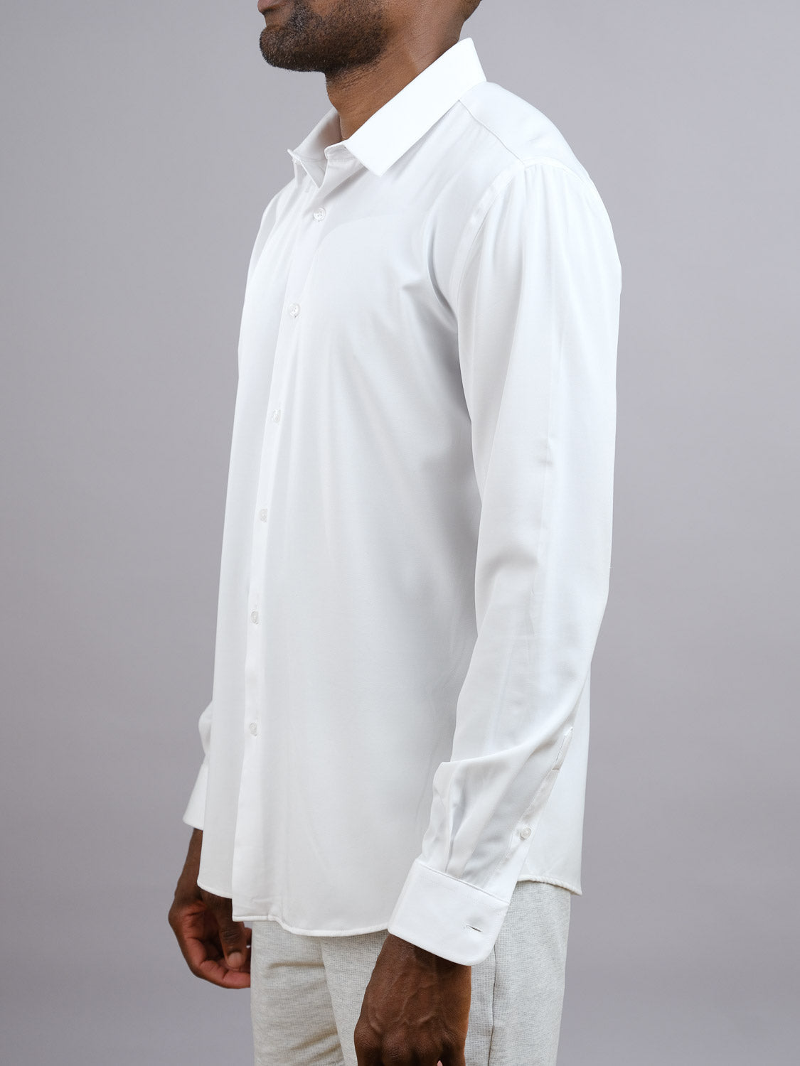 A man stands in profile against a plain light gray background, wearing the ID Menswear Alex 4-Way Stretch Slim Dress Shirt in white, paired with light-colored pants.