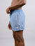 A man stands sideways against a plain light grey background, wearing ID Menswear’s Oyogu Quick Dry 4 Way Stretch Swim Trunk in light blue, made from recycled plastic bottles. His left arm is bent and resting near his waist.