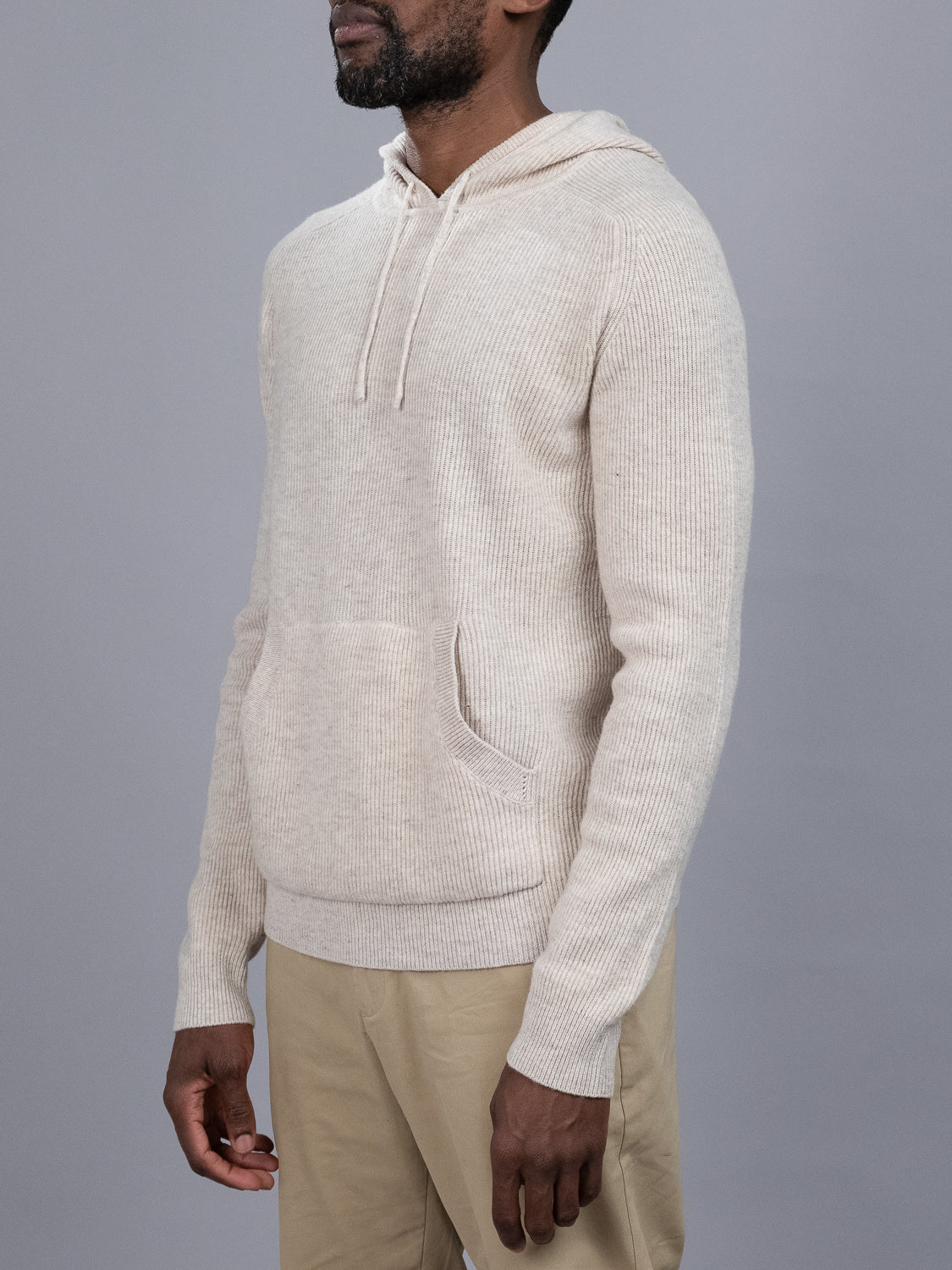 A man wears the Skien Merino Wool Hoody by ID Menswear—a light beige, luxurious knit with a front pocket—paired with khaki pants against a gray background, showcasing this refined, cold-weather essential.