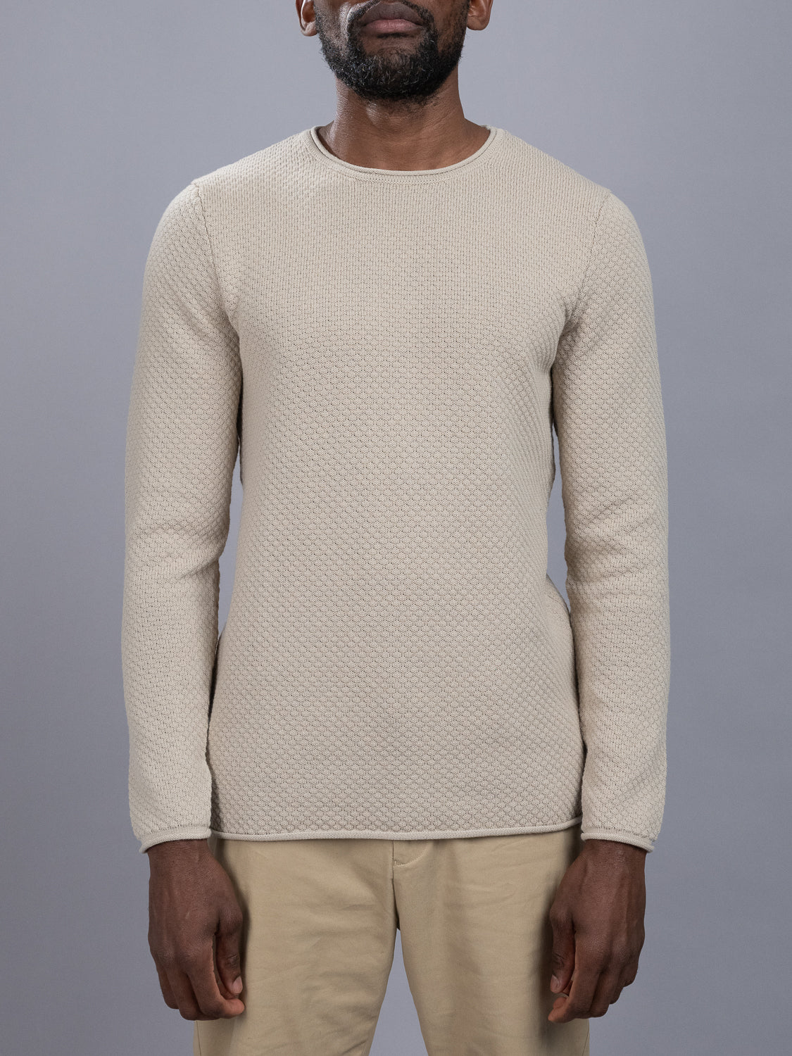 A person in a cream Westside Rolled Crew Neck Sweater with a subtle diamond pattern from ID Menswear and light khaki pants is against a plain gray backdrop. Their hands rest relaxed at their sides.