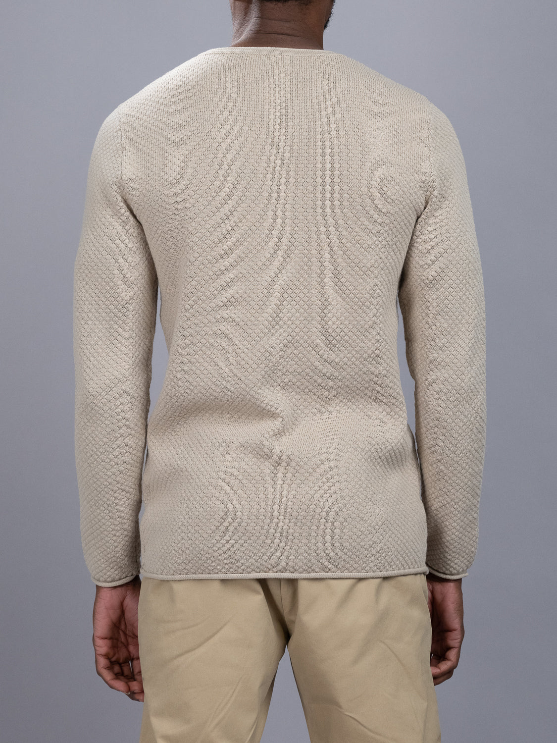 An individual displays the ID Menswear Westside Rolled Crew Neck Sweater from our knitwear collection, donning textured beige against a gray backdrop. The sweaters subtle pattern is paired with khaki pants as they stand with arms at their sides.