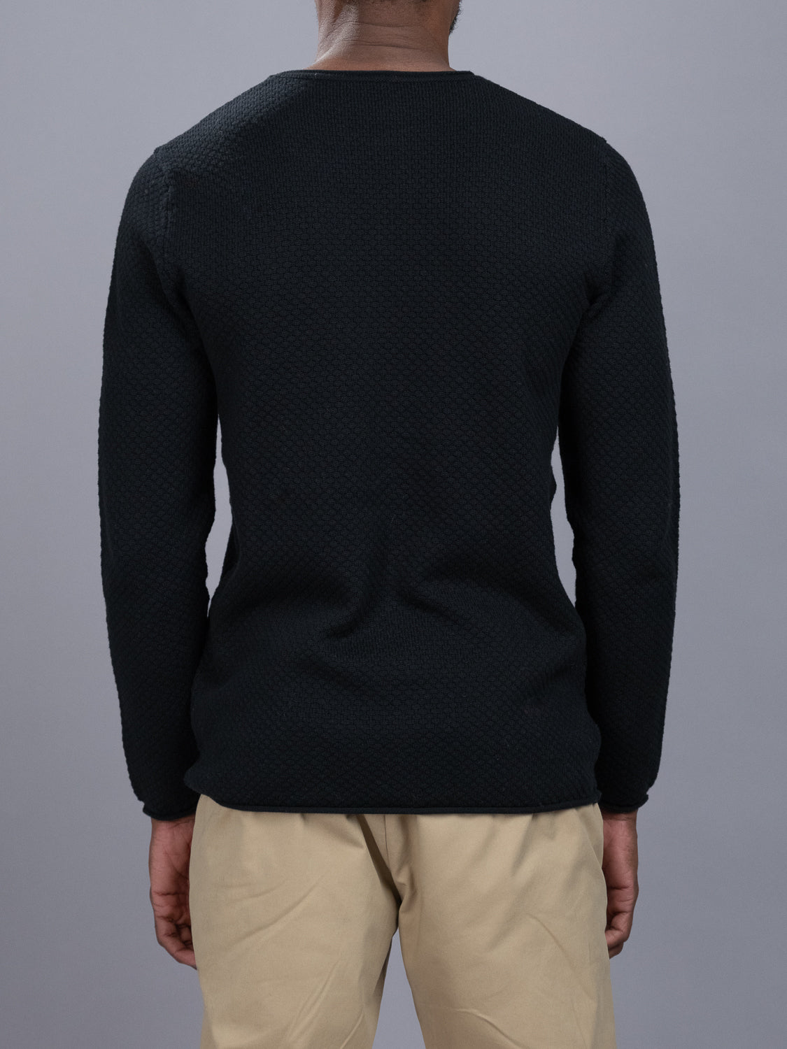 A person is seen from the back, wearing a black Westside Rolled Crew Neck Sweater by ID Menswear paired with beige pants against a plain gray background.