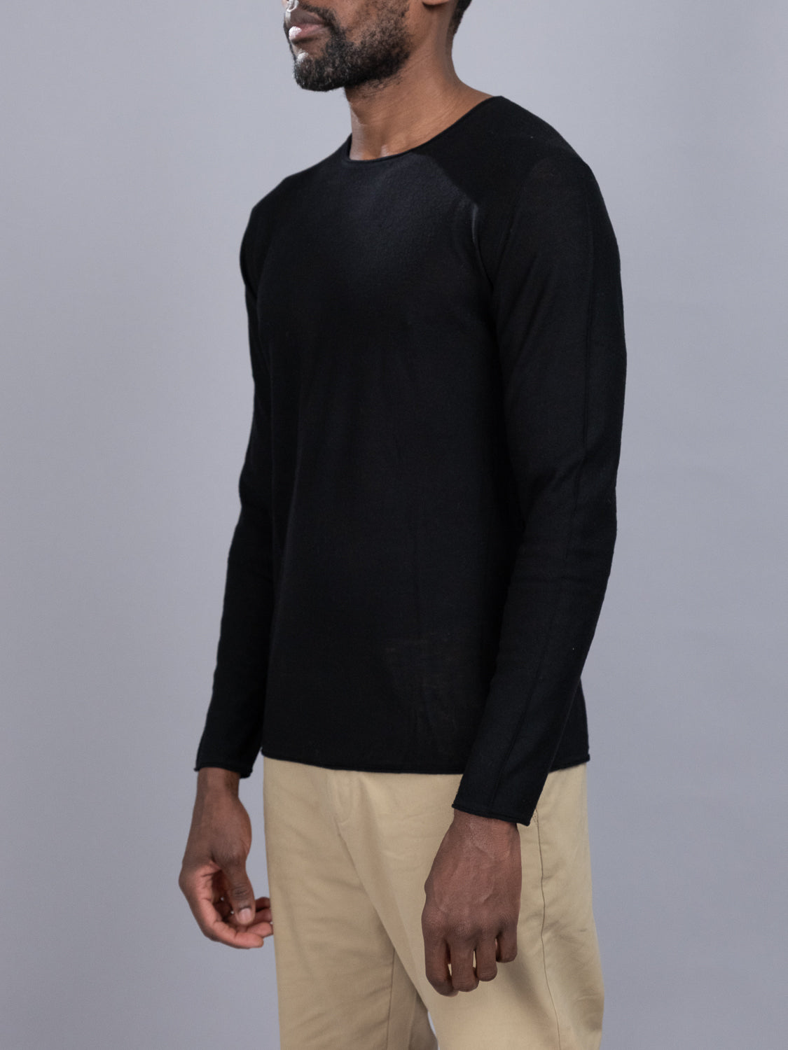 The individual is wearing a Vogue Lightweight Merino Sweater in black from ID Menswear and beige pants, standing against a plain gray background slightly facing right, with their face hidden.