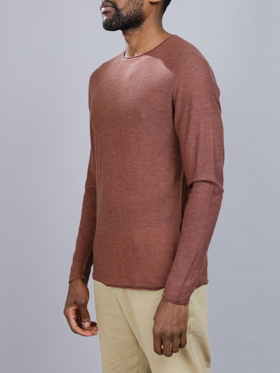 A person in a Vogue Lightweight Merino Sweater by ID Menswear, rust-colored and long-sleeved, paired with beige pants, stands against a gray background. Positioned sideways with their right arm slightly visible, they highlight the elegance of the reverse knit design.